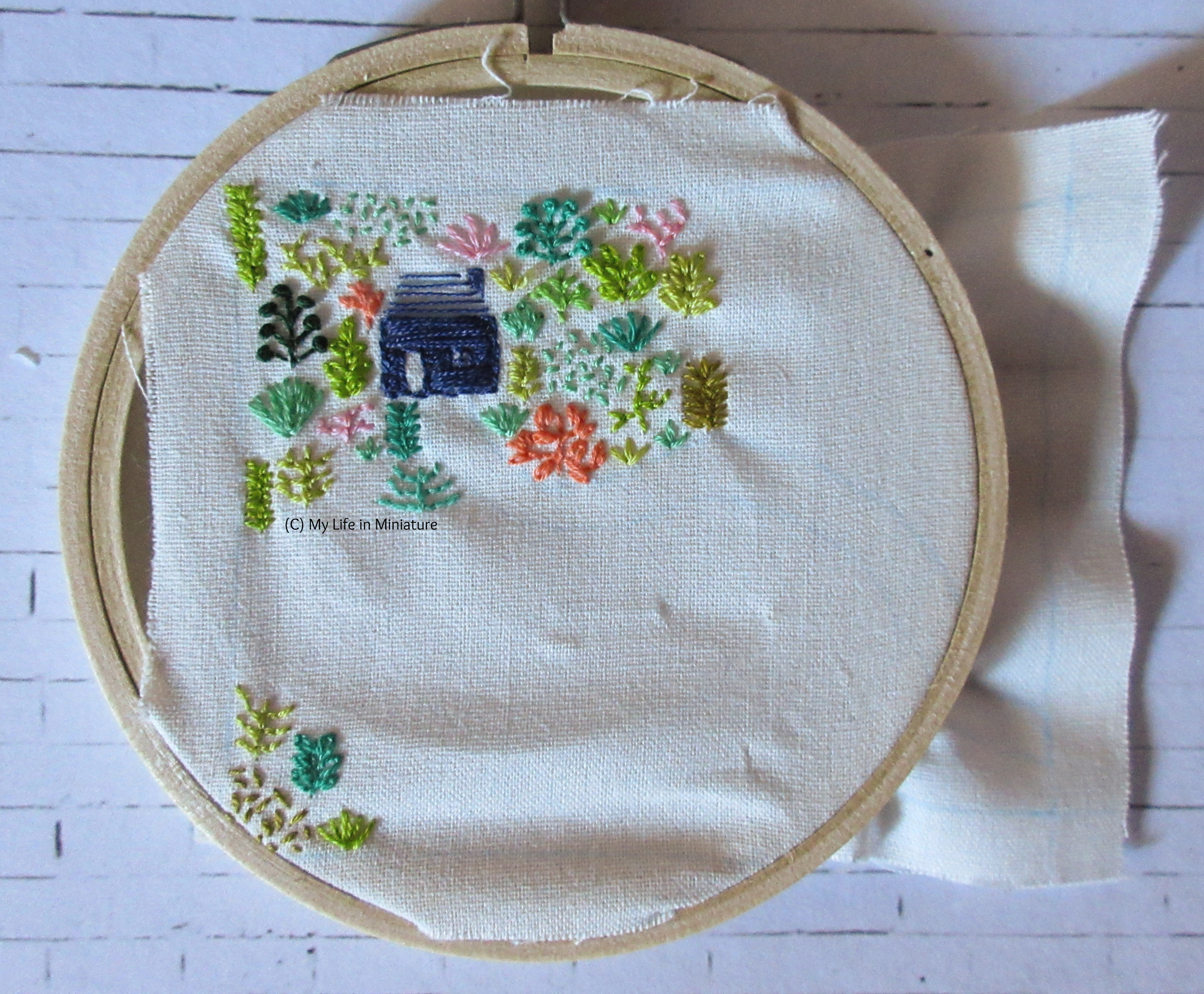 White fabric is in an embroidery hoop. Stitched on it is a small navy blue house, and an assortment of plants in different colours. Most of the plants and the house are in the top-left of the fabric, and the rest are in the bottom-left corner. 
