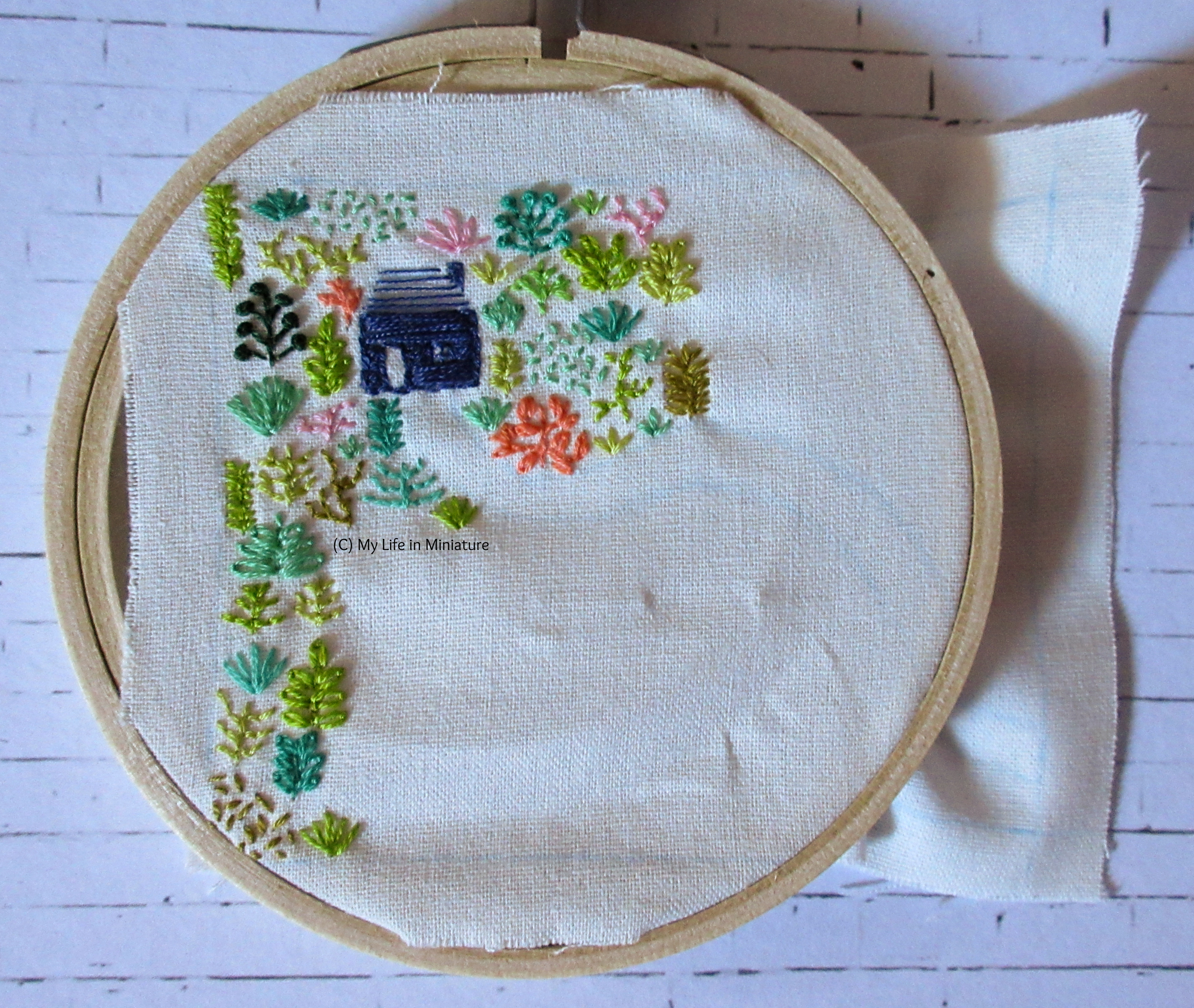 White fabric is in an embroidery hoop. Stitched on it is a small navy blue house, and an assortment of plants in different colours. All the stitching is towards the left-hand-side of the fabric. A white path is starting to emerge, curving from the front of the house. 