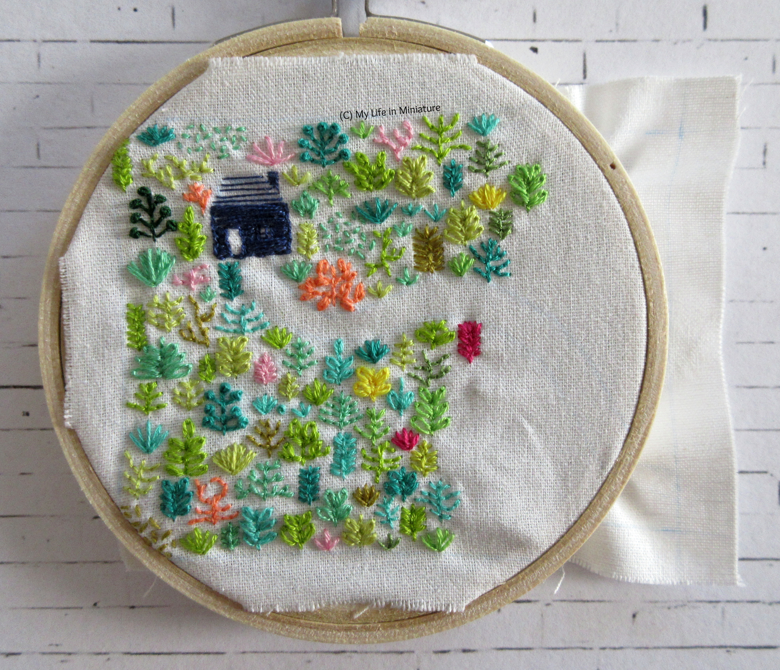White fabric is in an embroidery hoop. Stitched on it is a small navy blue house, and an assortment of plants in different colours. The plants cover most of the fabric in the hoop, and a winding path leading from the house is visible.