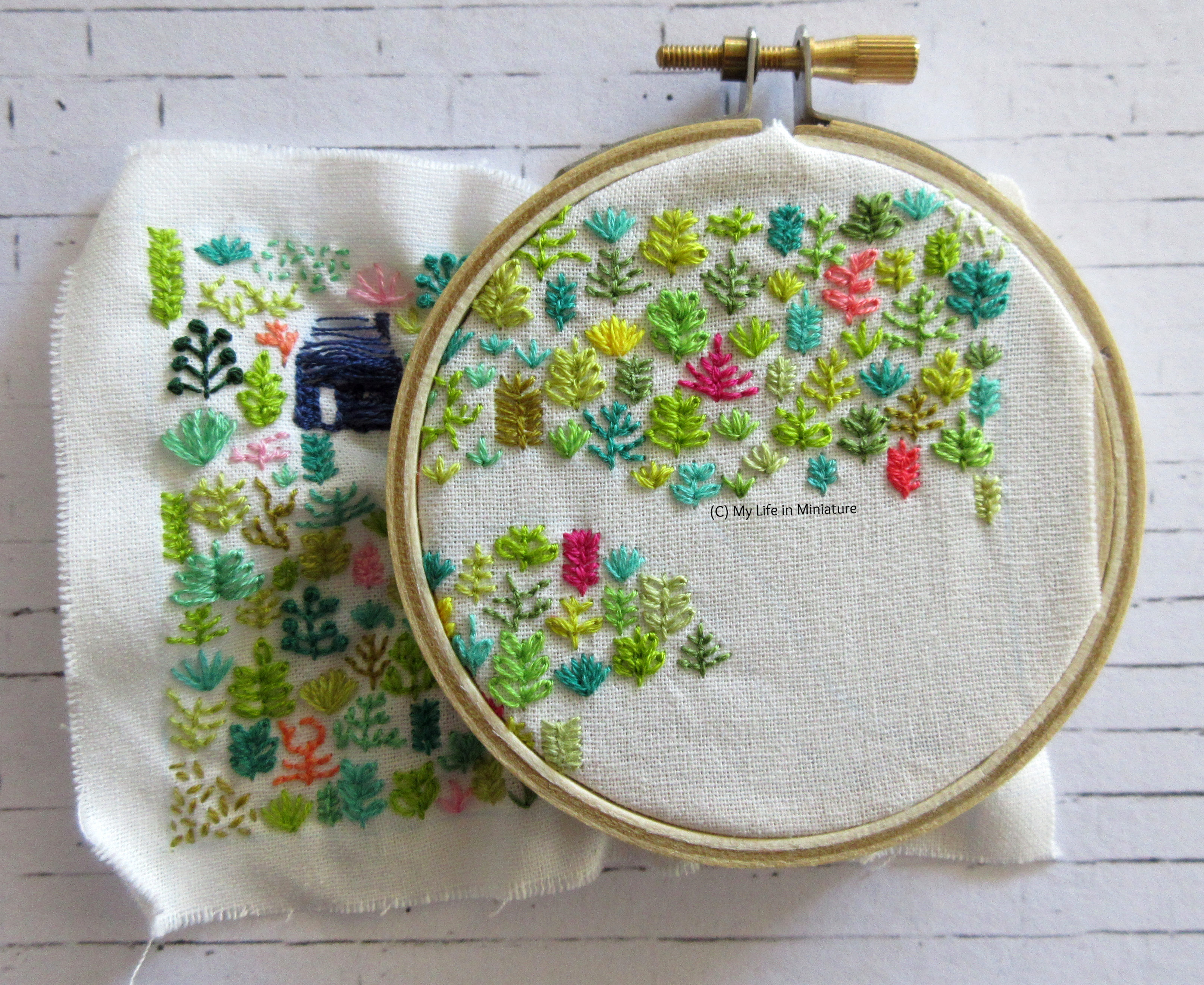 White fabric is in an embroidery hoop. Stitched on it is a small navy blue house, and an assortment of plants in different colours. The hoop is in the top-right of the fabric, and is about half-full of plants.