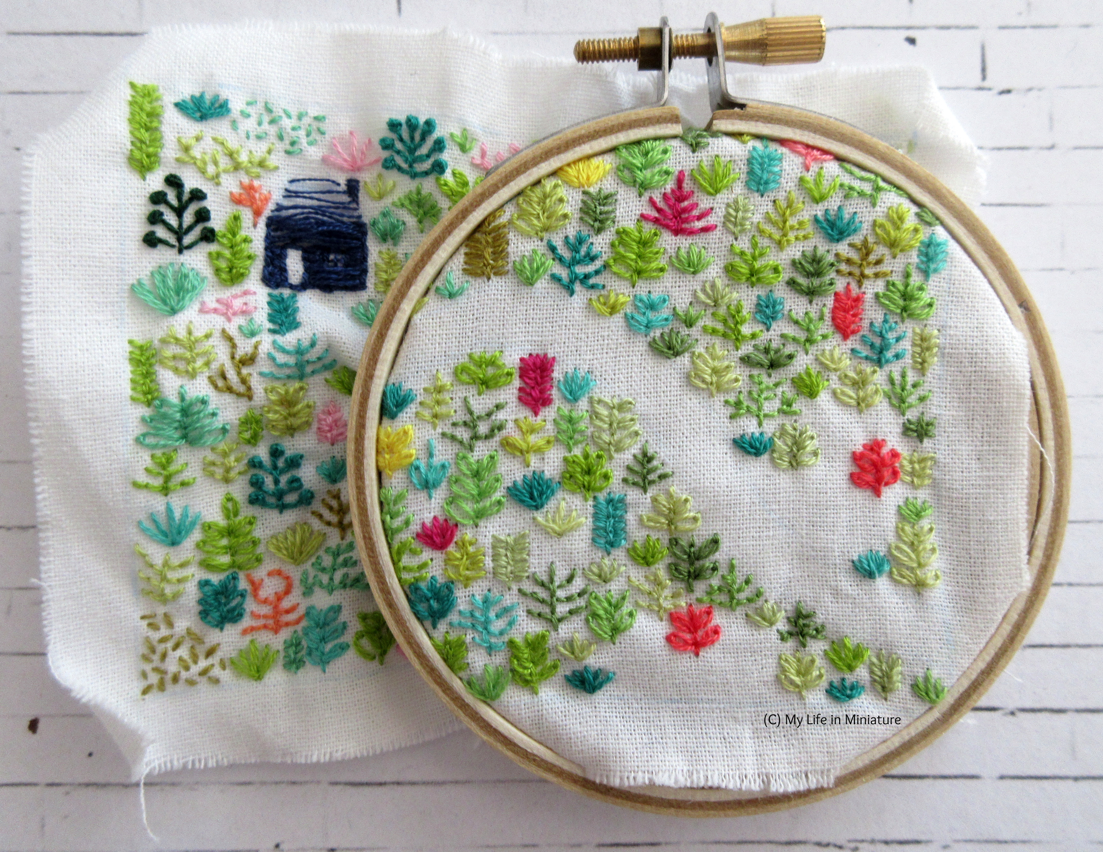 White fabric is in an embroidery hoop. Stitched on it is a small navy blue house, and an assortment of plants in different colours. The hoop is in the top-right of the fabric, and is nearly full of plants.