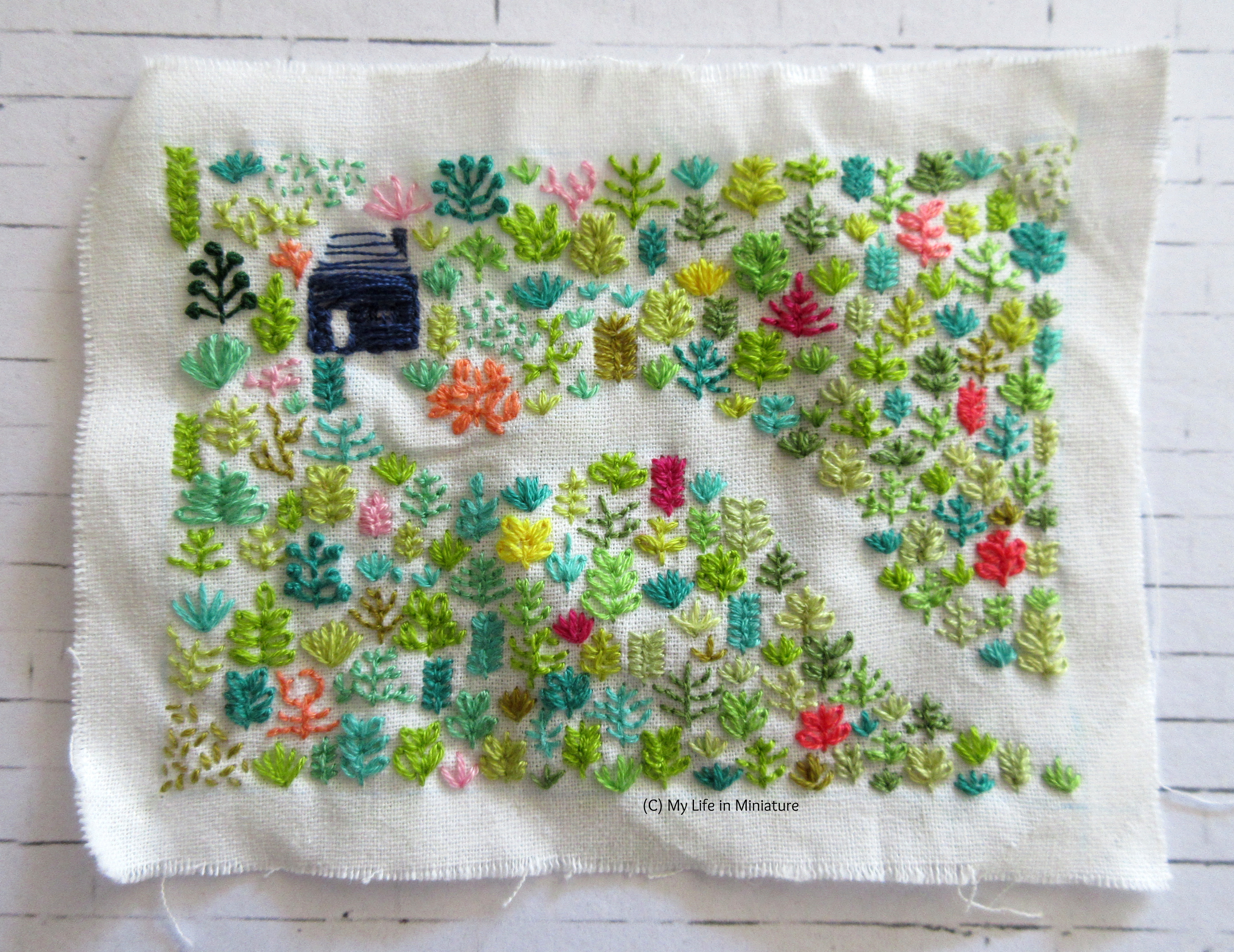 A square of white fabric sits on a white background. Embroidered on it is a navy blue house, surrounded by plants that also line a path, curving towards the bottom-right.