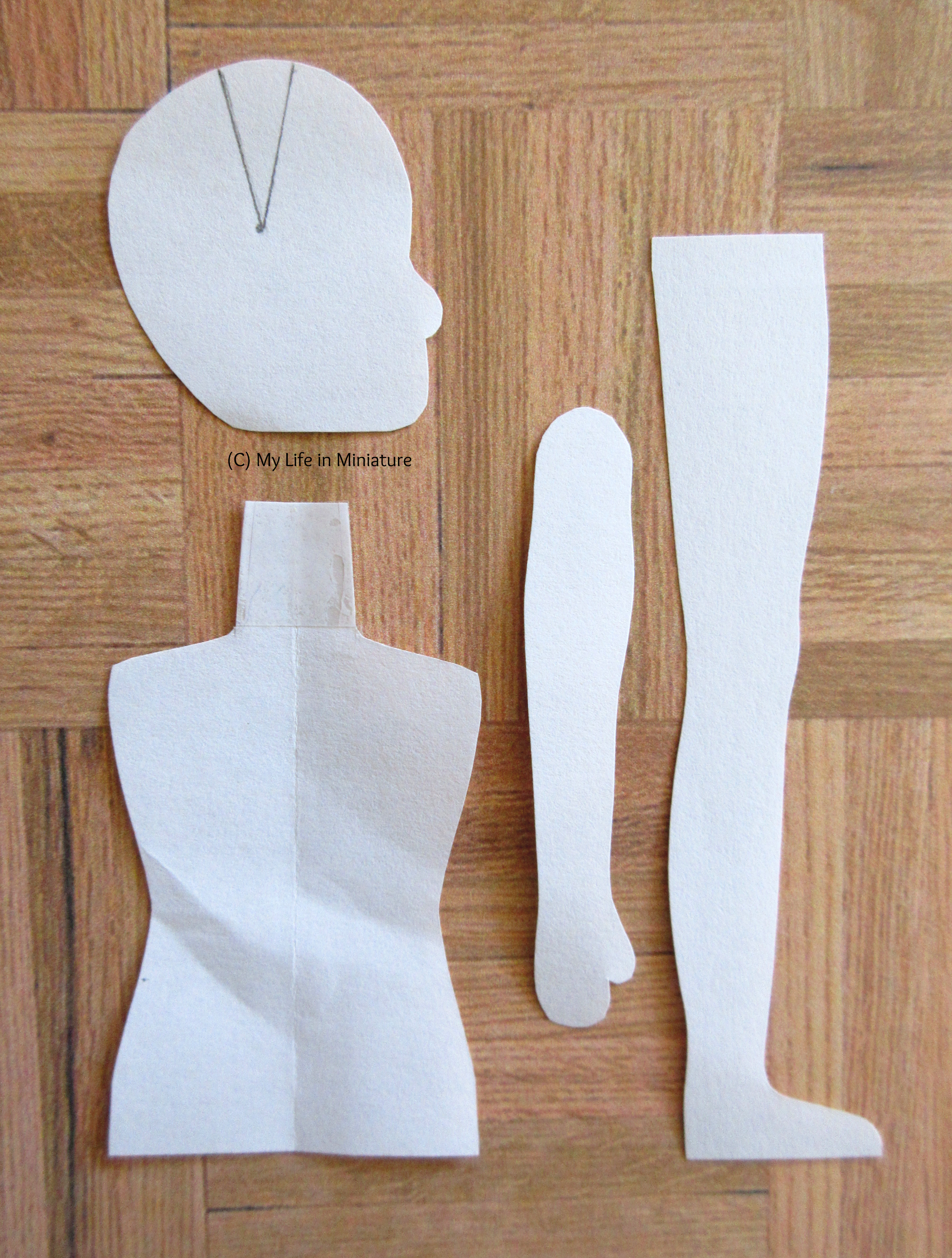 A paper pattern for a doll sits on a wood parquet background. Pieces for a head, arm, leg, and torso are visible.