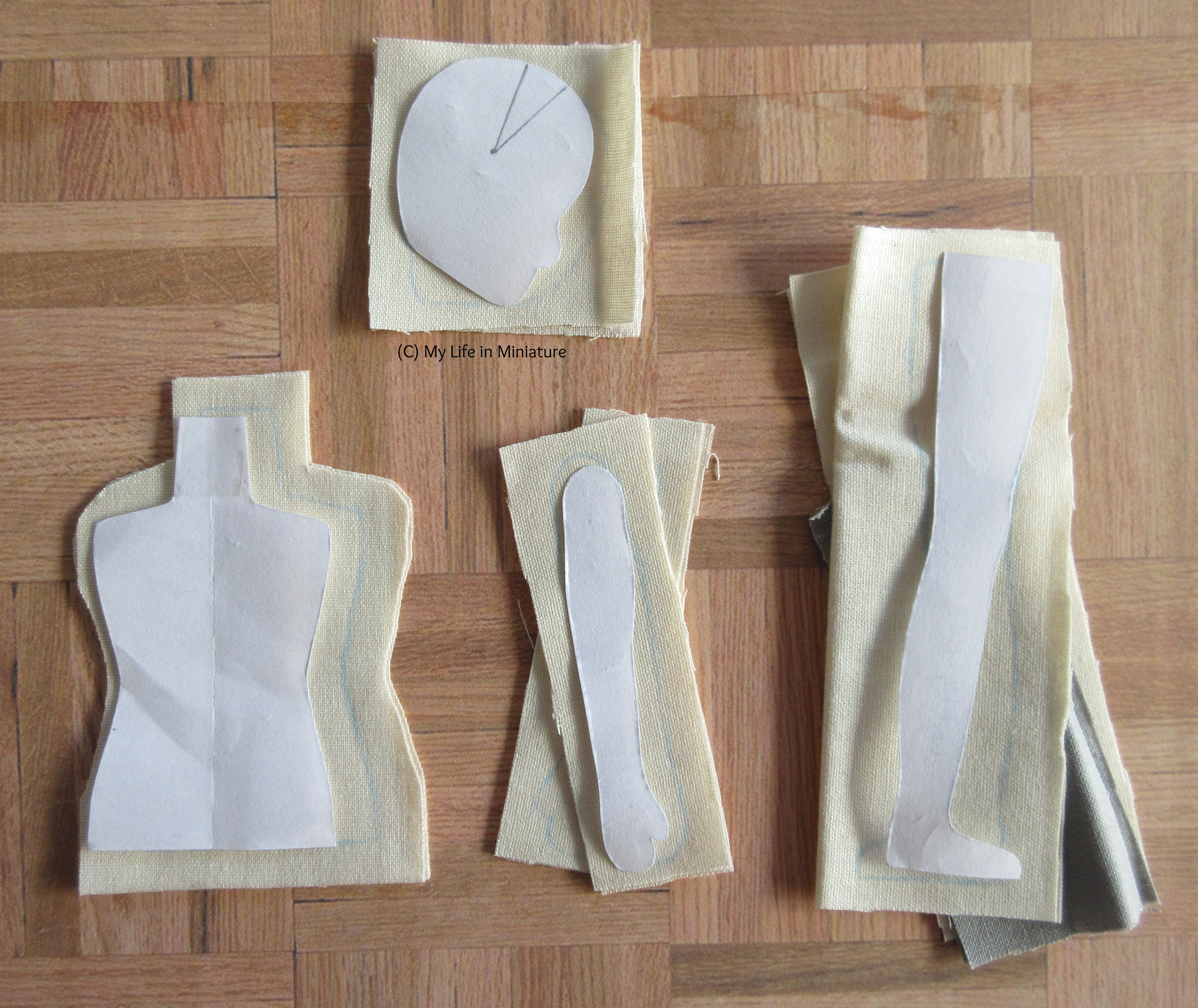 Paper pattern pieces for a doll, with the fabric they're traced onto with blue chalk, sit on a wood parquet background.