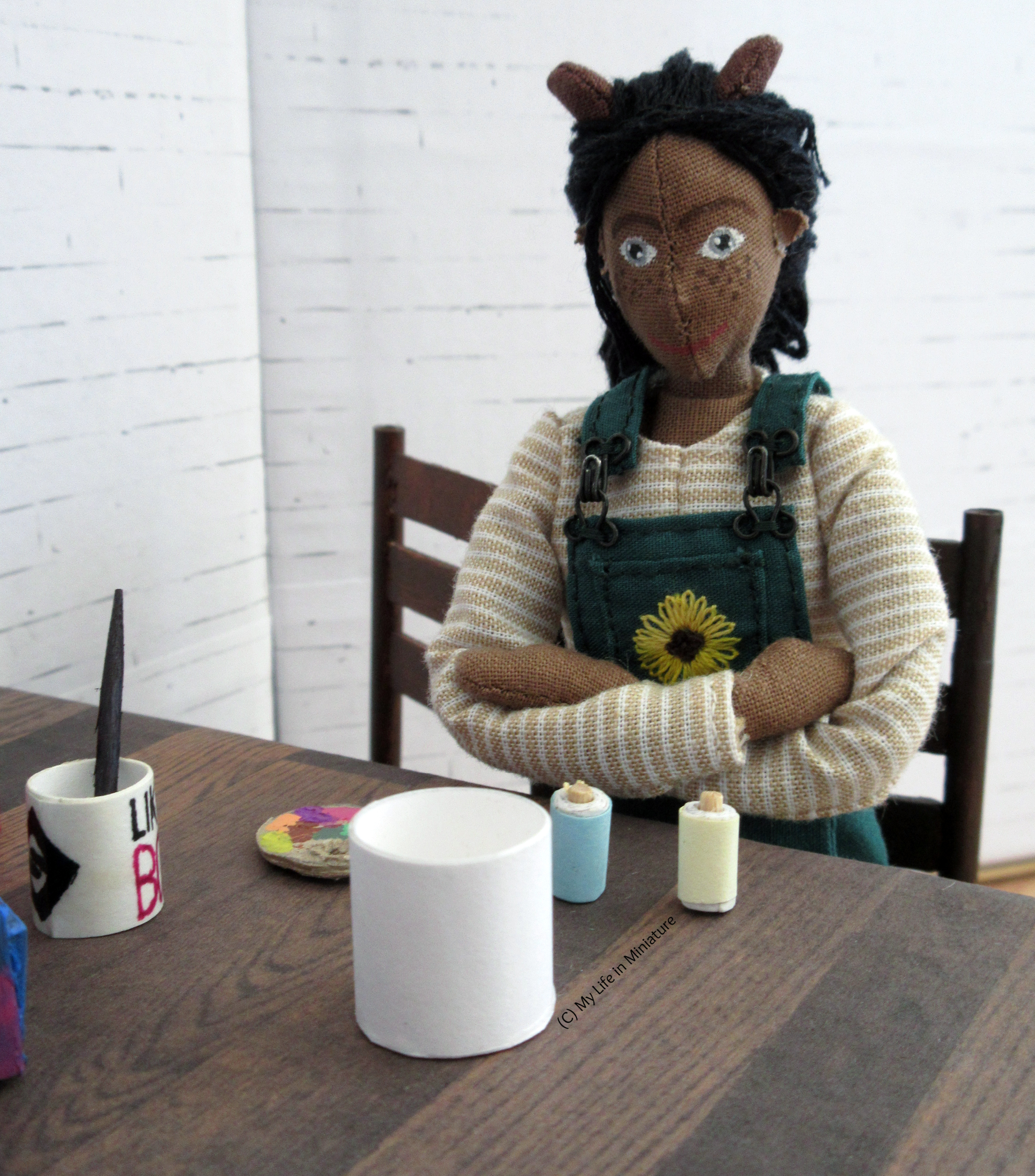 Petra sits at a table, painting supplies set up. She has crossed arms and is looking between a pale blue paint tube and a yellow paint tube. A white pot also sits in front of her.