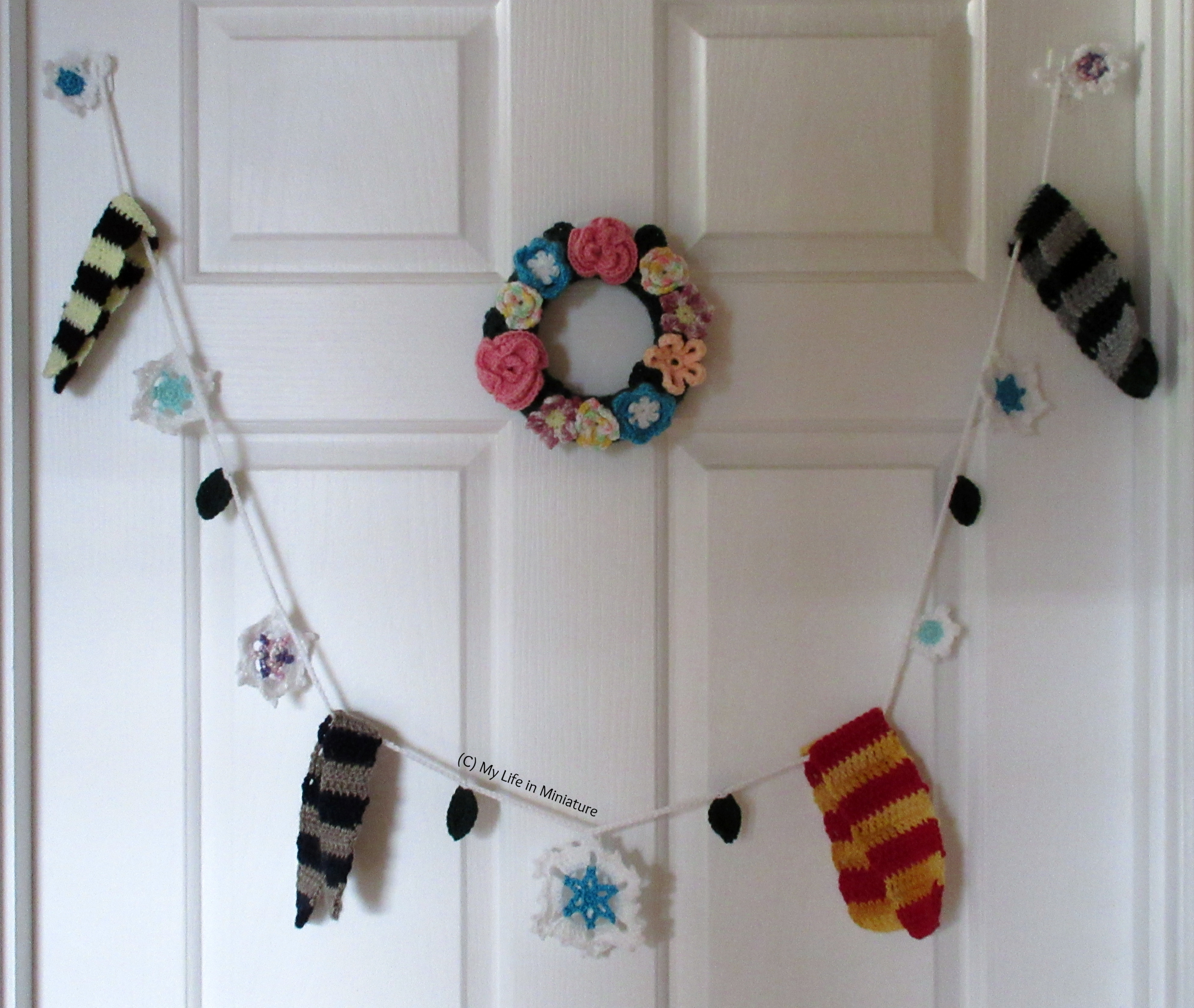 A crocheted garland, made of scarves, snowflakes, and some leaves, hangs on a white door below a small floral crocheted wreath.