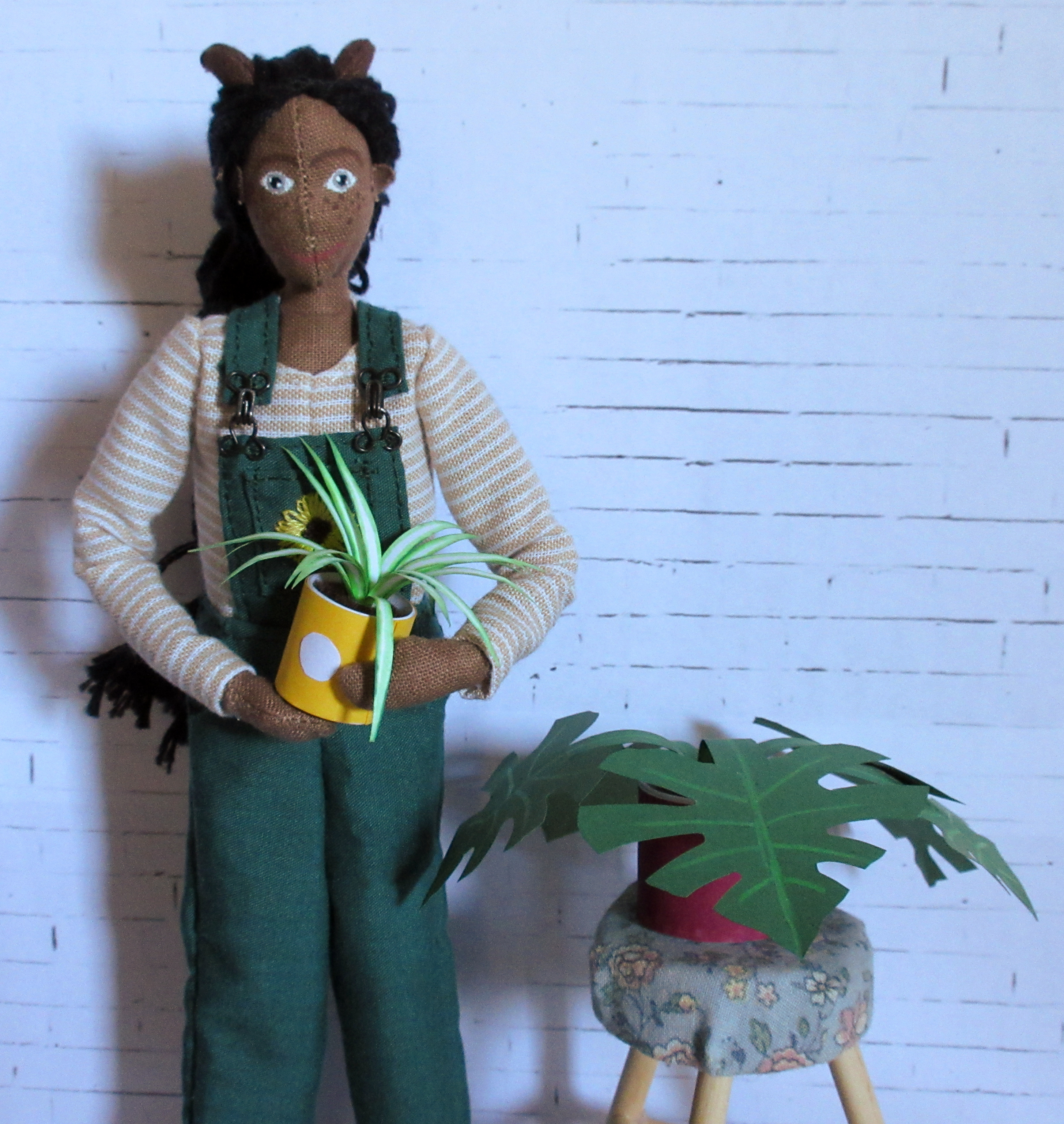 Petra stands next to a stool, on which is the paper monstera. She holds her spider-plant in her arms, and smiles at the camera.