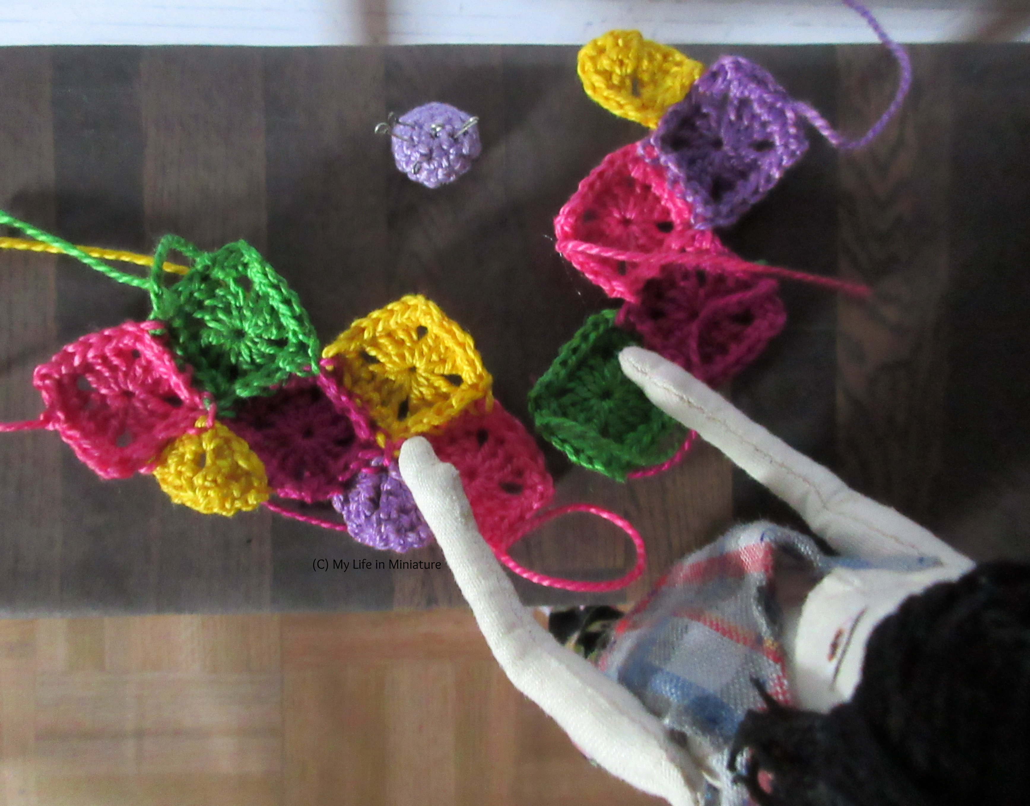 Tiffany stands over a table, on which is an assortment of different-coloured crocheted squares. She's sewing two of them together.