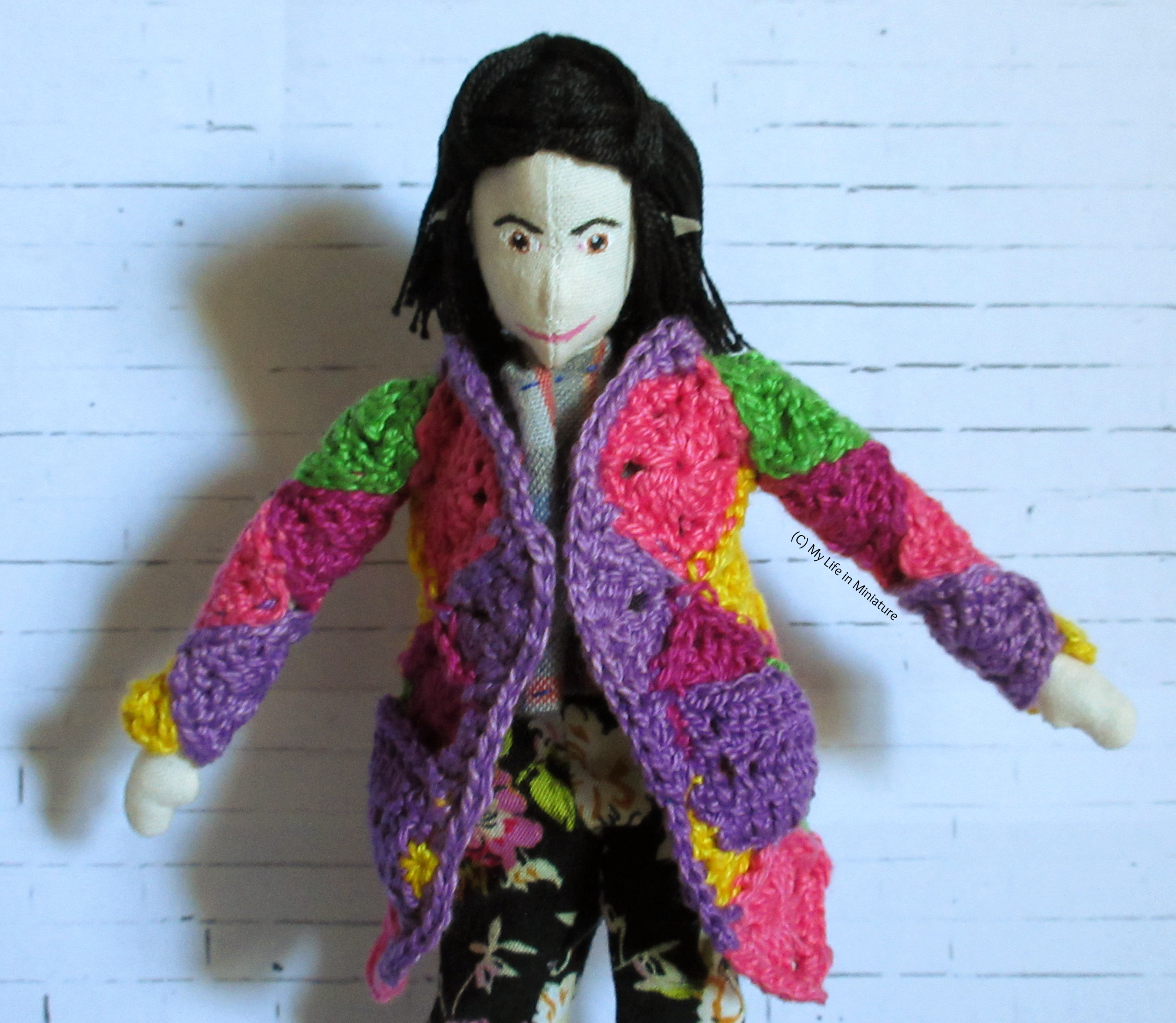 Tiffany wears a colourful crocheted cardigan against a white brick background. The cardigan is made of crocheted squares in purple, light and dark pink, green, and yellow.