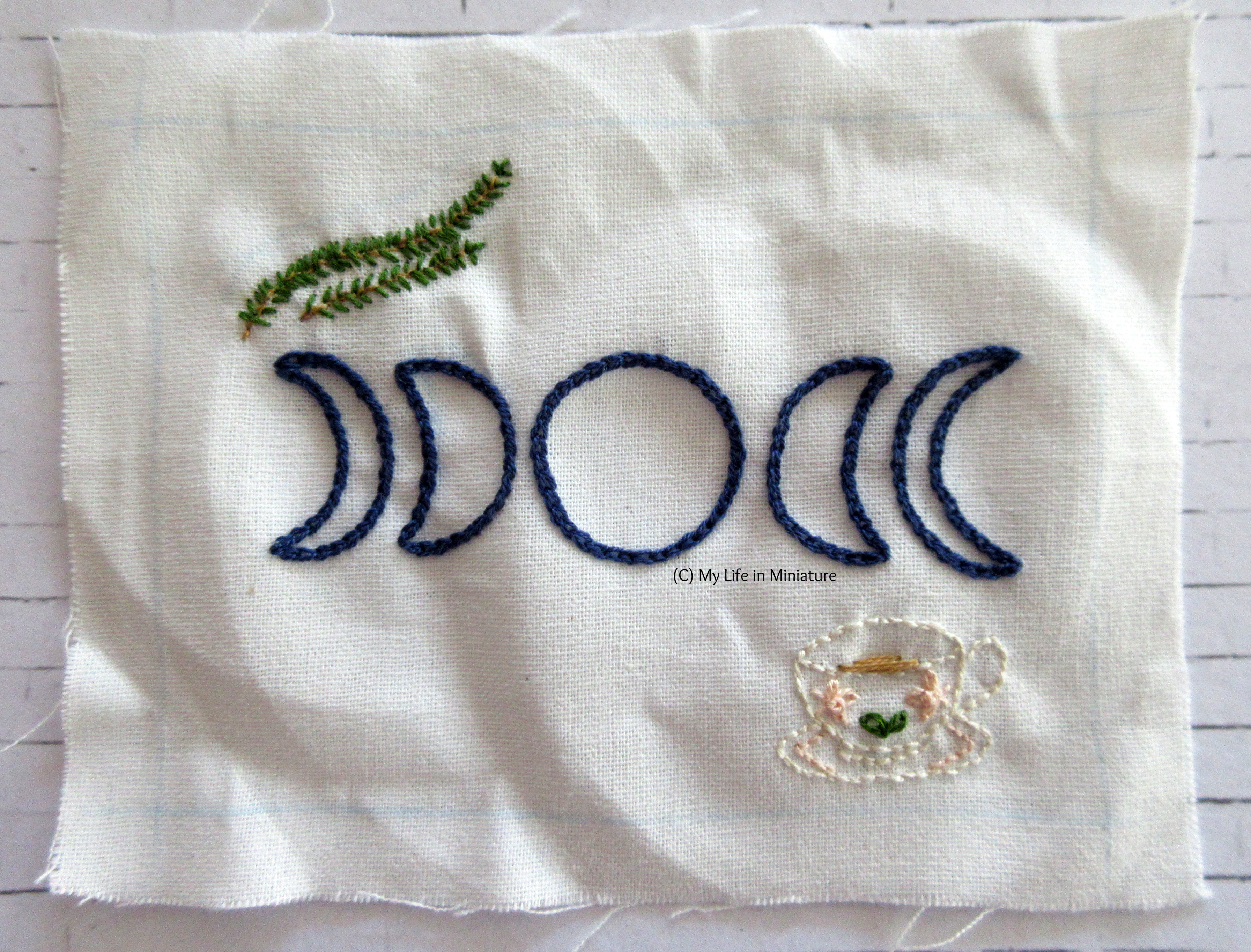 A square of white fabric sits on a white brick background, with five moons embroidered on in navy blue. Two sprigs of rosemary are in the top-left, and a cream teapot with pink flowers and leaves on is in the bottom-right. There is also a little bit of tea in the teacup.