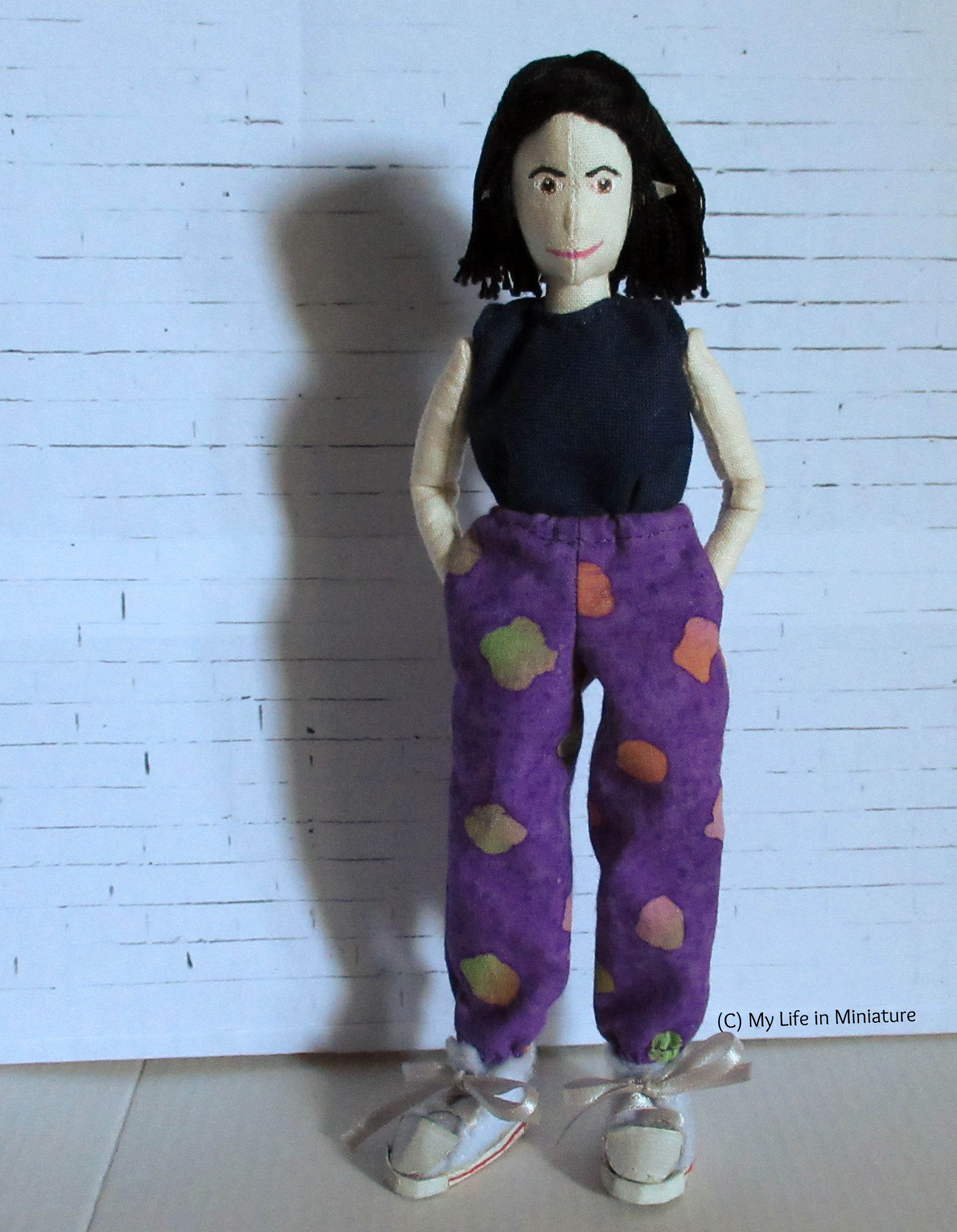 Tiffany stands before a white brick background. She wears loose pants that have elastic gathering at the waist and ankles, hands in pockets. The fabric is purple and batik-printed, and she wears it with a navy top. She looks at the camera. 