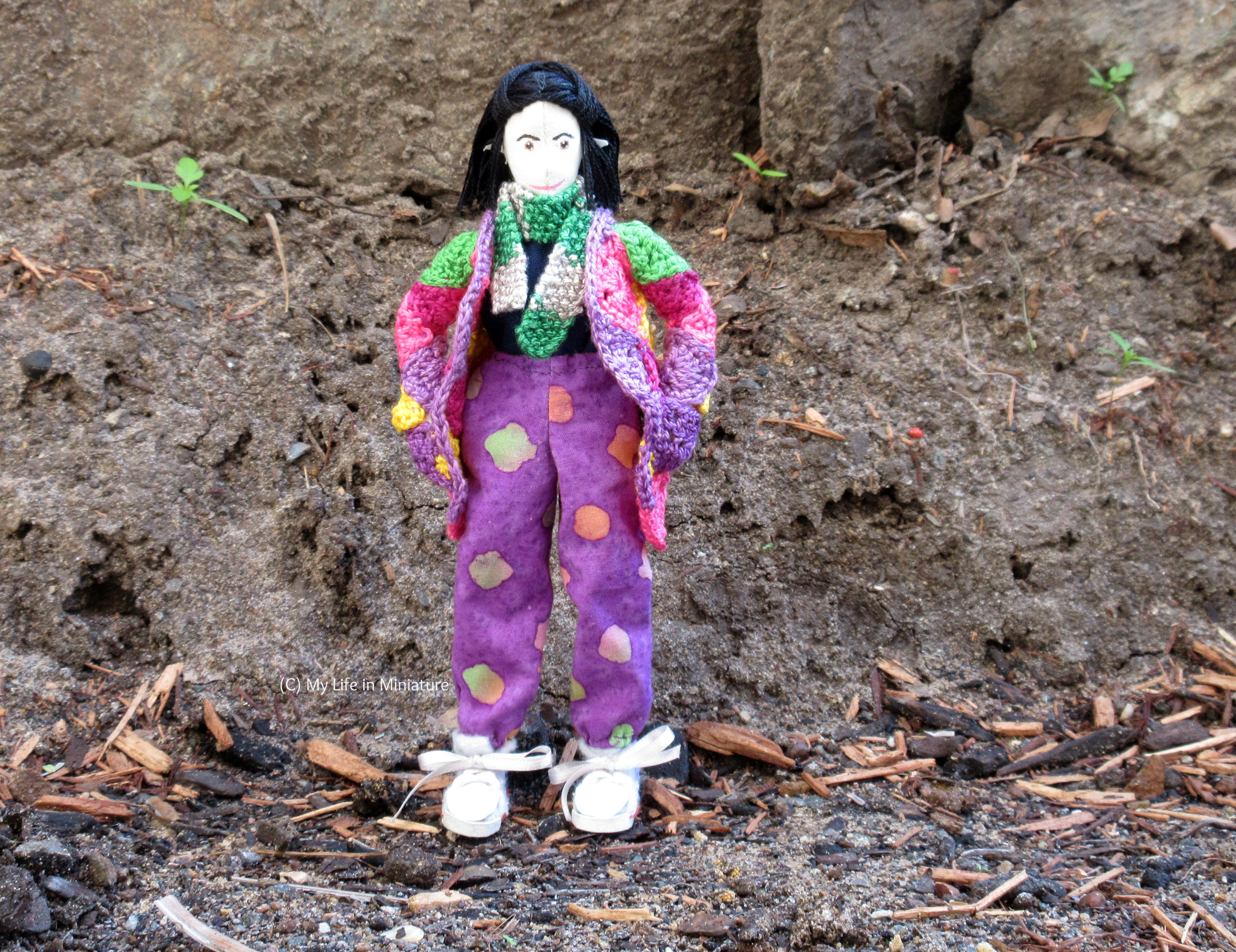 Tiffany stands outside, against a dirt wall. She wears the purple, loose pants with a navy top, her crocheted cardigan, and a scarf. She looks at the camera, hands in cardigan pockets.