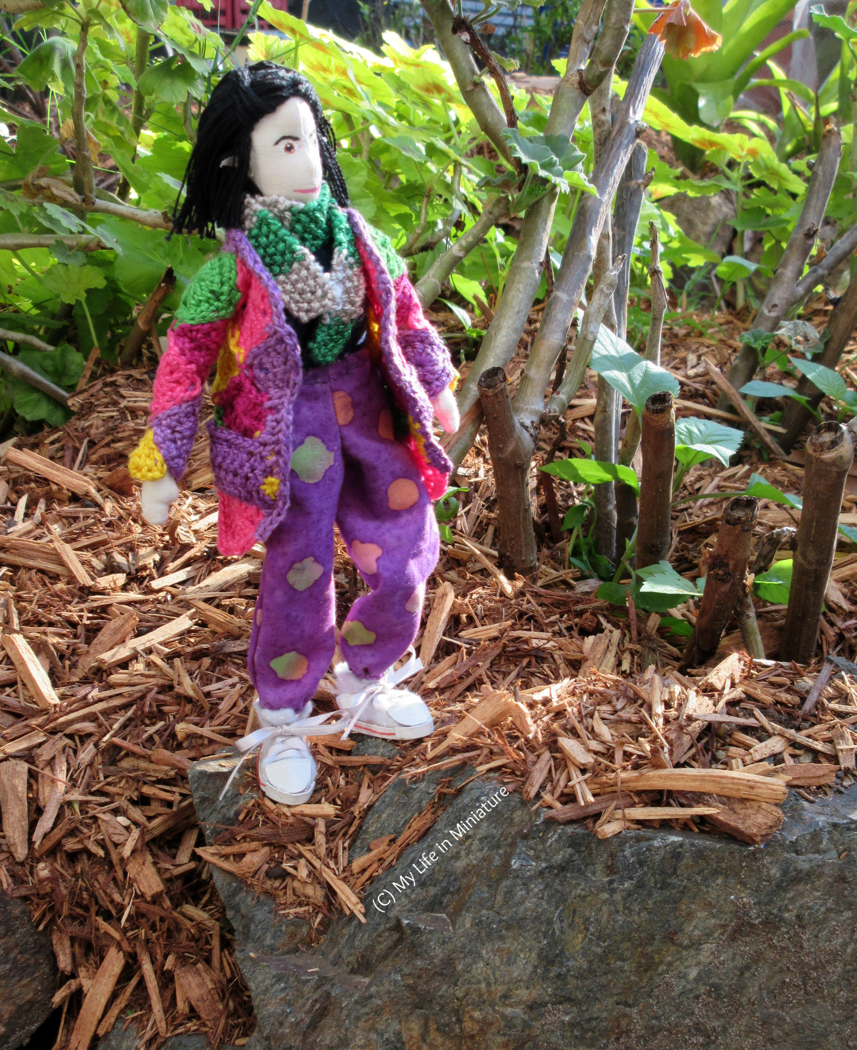 Tiffany stands upon a rock with plants in the background, looking heroically up to the sky. She wears the loose purple pants, crocheted cardigan, and scarf.