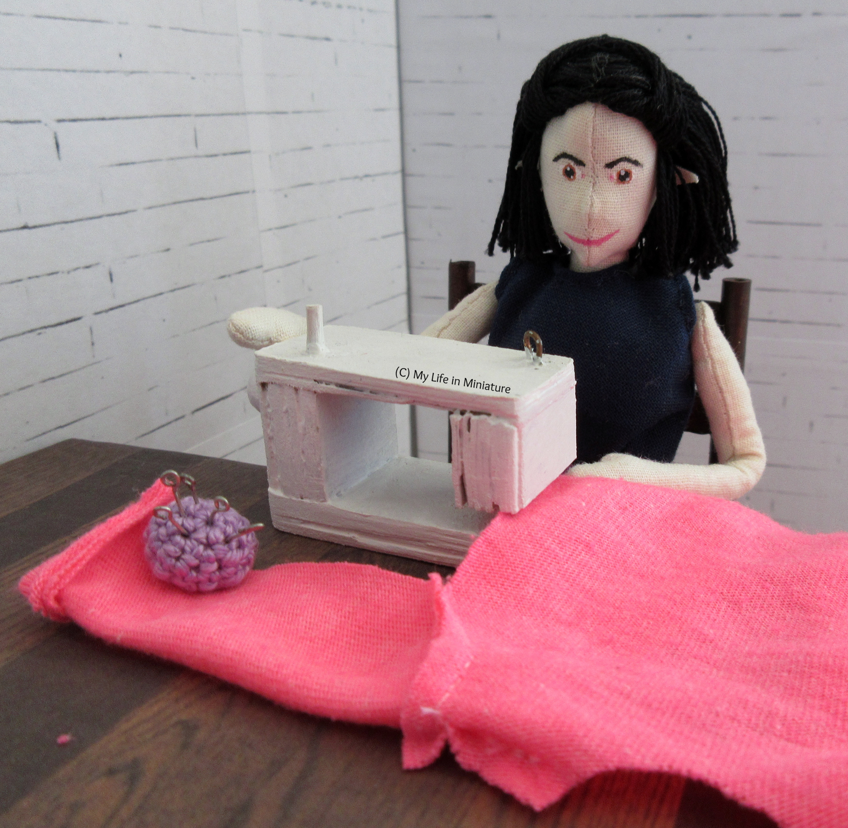 Tiffany sits at her sewing machine, sewing a side seam of the neon pink cardigan. A cuffed sleeve is visible, with a purple pincushion resting on it.