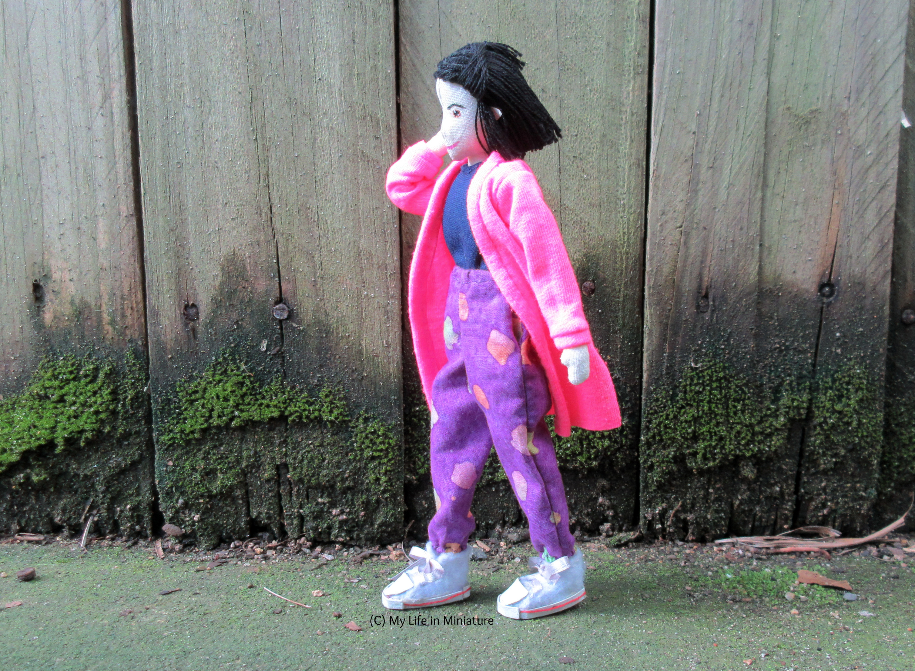 Tiffany walks left-to-right in front of a wood fence. She wears the neon pink cardigan with her loose purple pants and a navy top, ends flowing in the breeze. She tucks some loose hairs behind her ear.