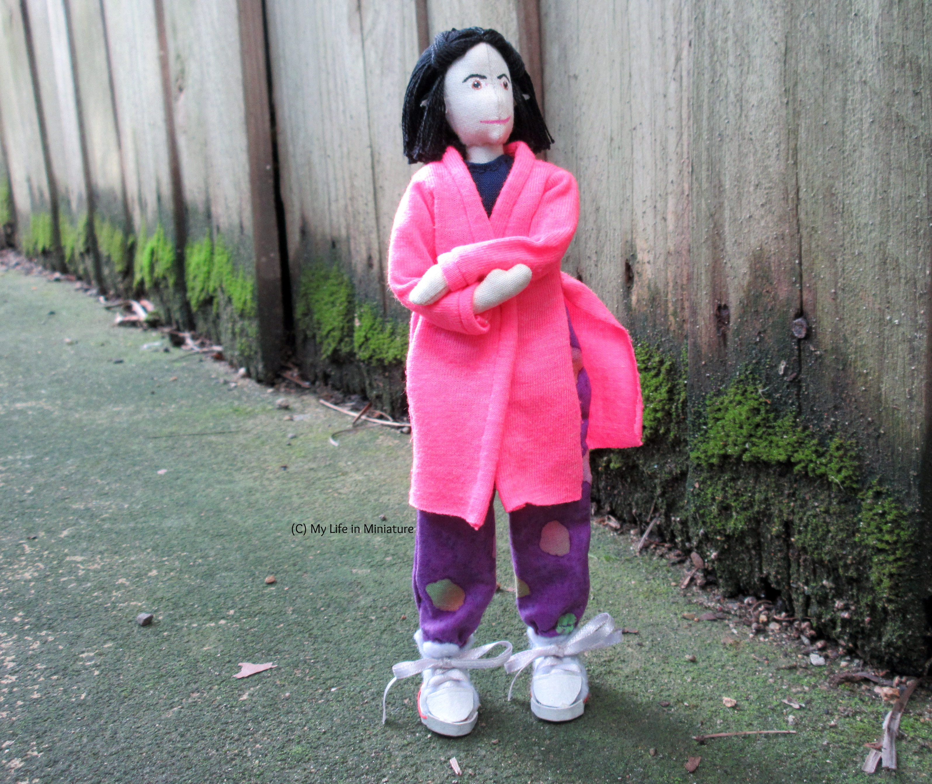 Tiffany stands near a wood fence, arms crossed and looking up annoyedly. She wears her neon pink cardigan with her loose purple pants and a navy top, ends flapping in the breeze.