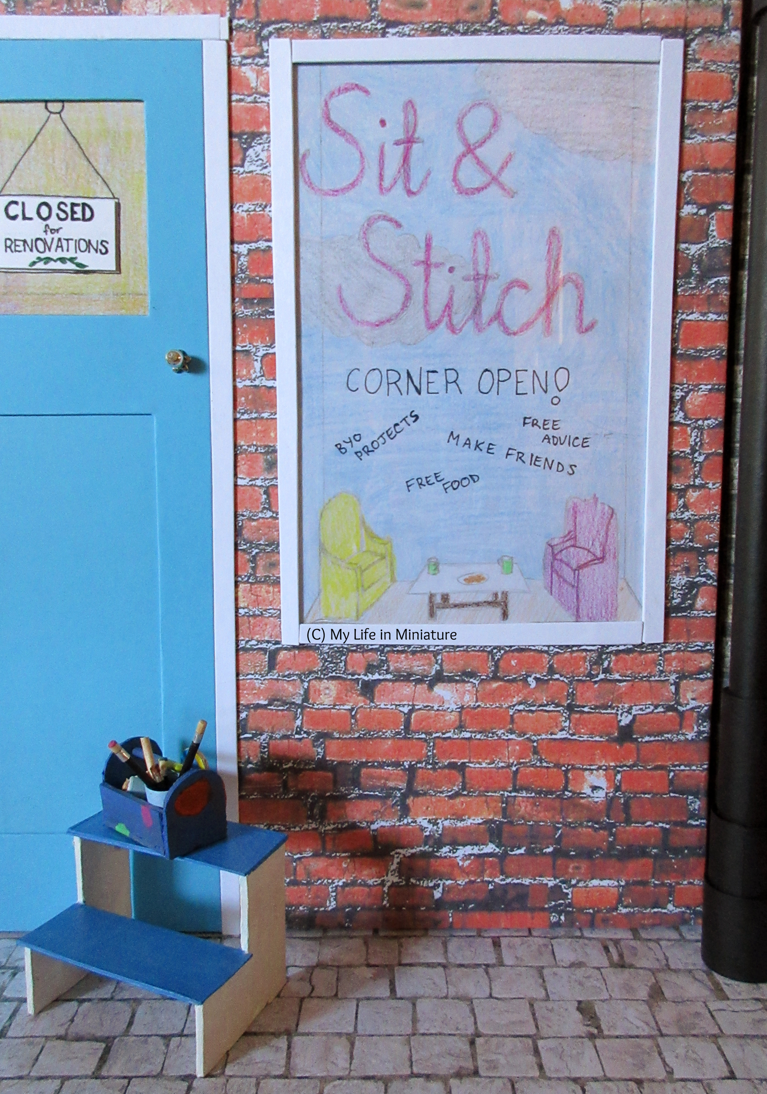 Clear shot of the 'Sit & Stitch' window display. The background to the display is blue sky with some silver clouds; the writing is purple and on the window. At the bottom is a miniature seating area, along with text that says 'Corner Open!' and some of the things offered. 