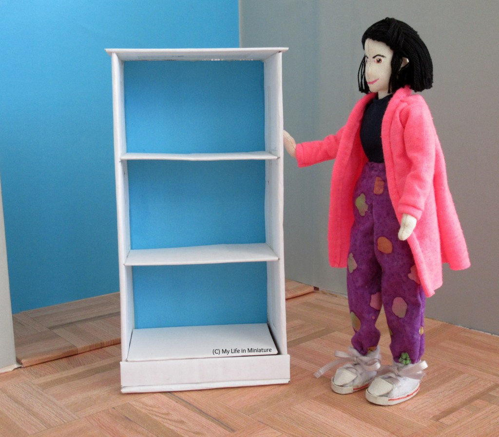Tiffany stands beside a white bookshelf that is about her height. It has three shelves and is bright blue on its back panel.