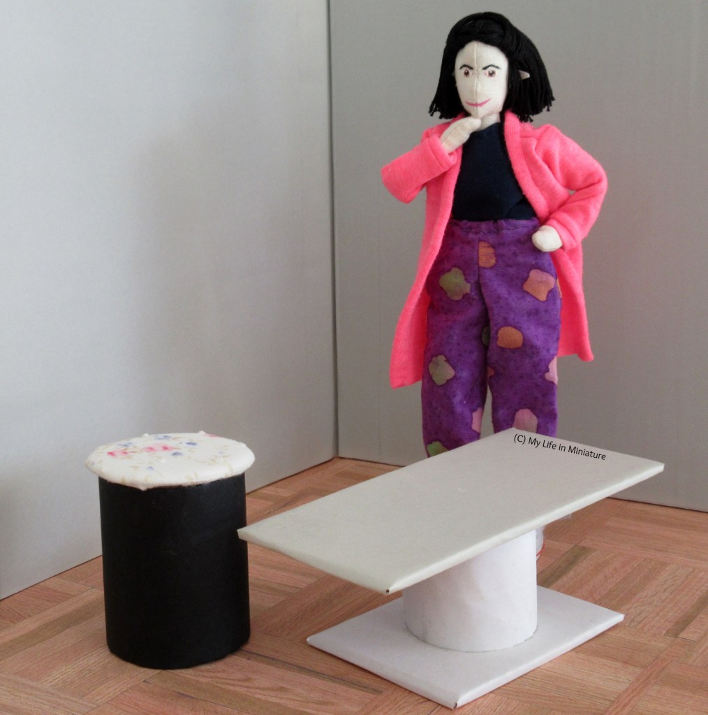 Tiffany stands with hand on hip, chin in hand, thinking. In front of her is a small coffee table (white base and grey top), and a stool (black base, white floral top). 