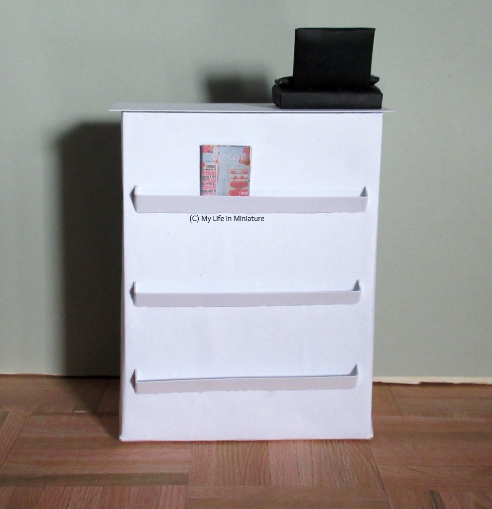 Front view of the Needle & Thread counter. It's white; a black cash register sits on the top right. There are three magazine shelves down the front; the top one has a craft magazine in.