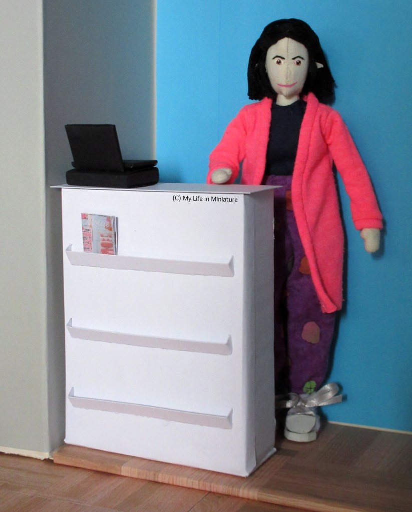 Tiffany stands behind the counter, hand resting on its surface. The cash register sits on top, and a craft magazine in the top magazine rack.