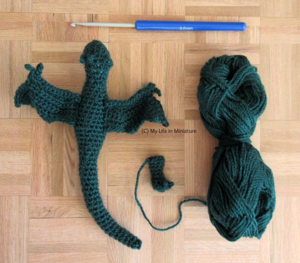 Flatlay of a 3mm crochet hook, a ball of dark green yarn, a dark green crocheted dragon shape without legs, and a small dark green crocheted foot with claws.