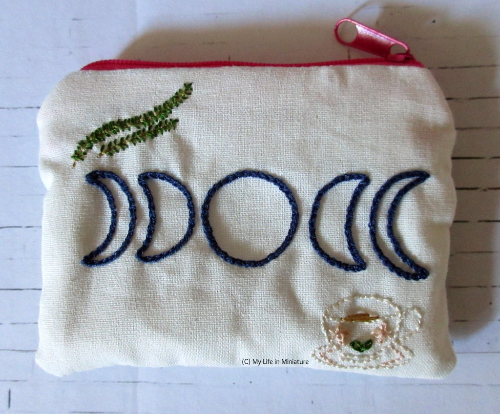 Flatlay view of other side of white cotton zippered pouch, embroidered with five navy moon phases, two rosemary stems, and a cup of tea. The zip is bright pink.