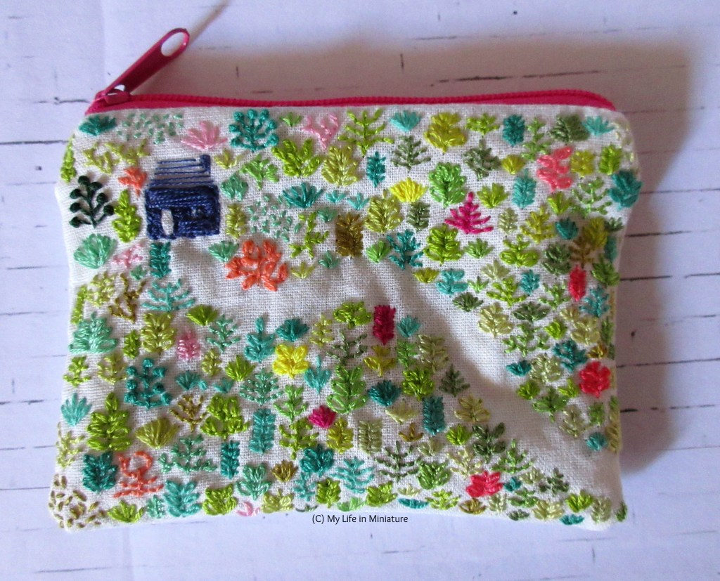 Flatlay view of a white cotton zippered pouch, embroidered with a navy blue house in a green forest. A path through the forest is created with negative space. The zip is bright pink.