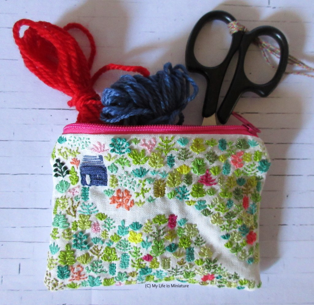 Flatlay view of the zippered pouch, forested side up. Spilling out of it is a pair of black embroidery scissors, and two small skeins of yarn: one red, one blue.