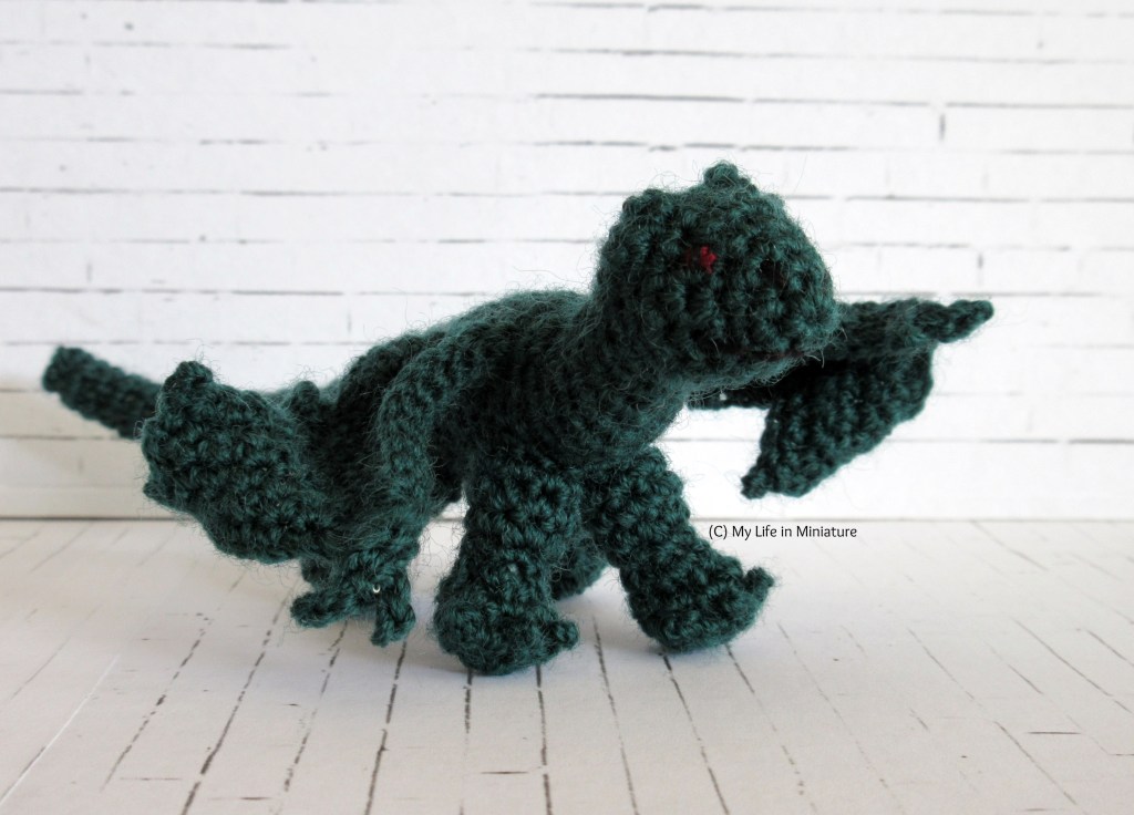 A dark green crocheted dragon stands in front of a white background.