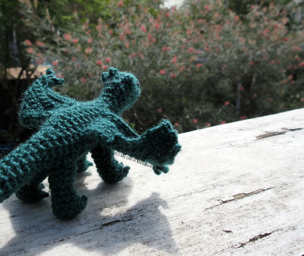 The dragon sits on a weather-damaged white wood board, looking out and down over a tree with red flowers.