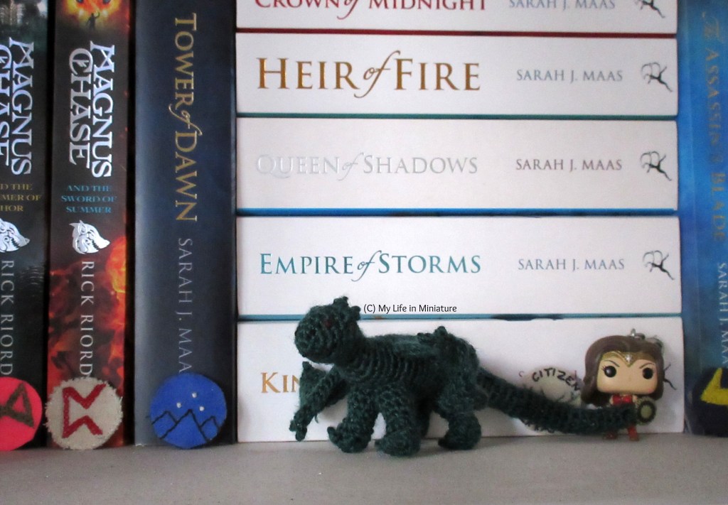 The dragon sits in front of various fantasy novels on a white bookshelf.
