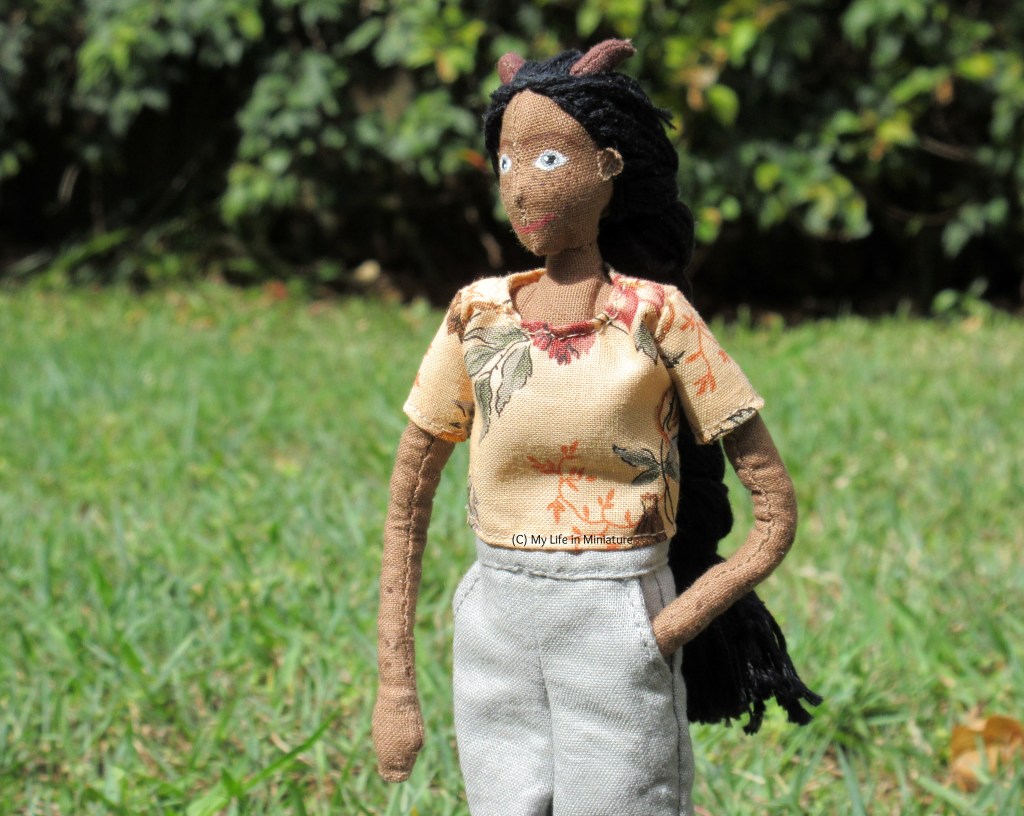 Petra walks on a lawn in the full sun, a green hedge behind her. She looks to the left and has a hand in her pocket.