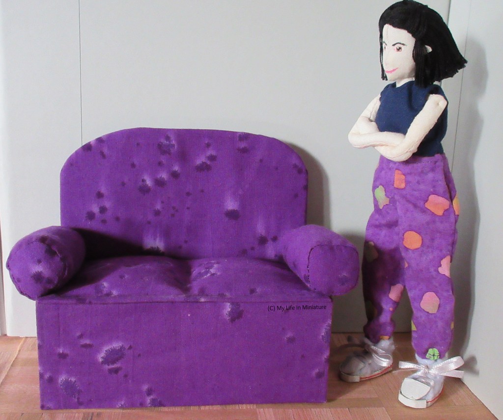 Tiffany stands to the side of the purple couch, arms folded in thought. 