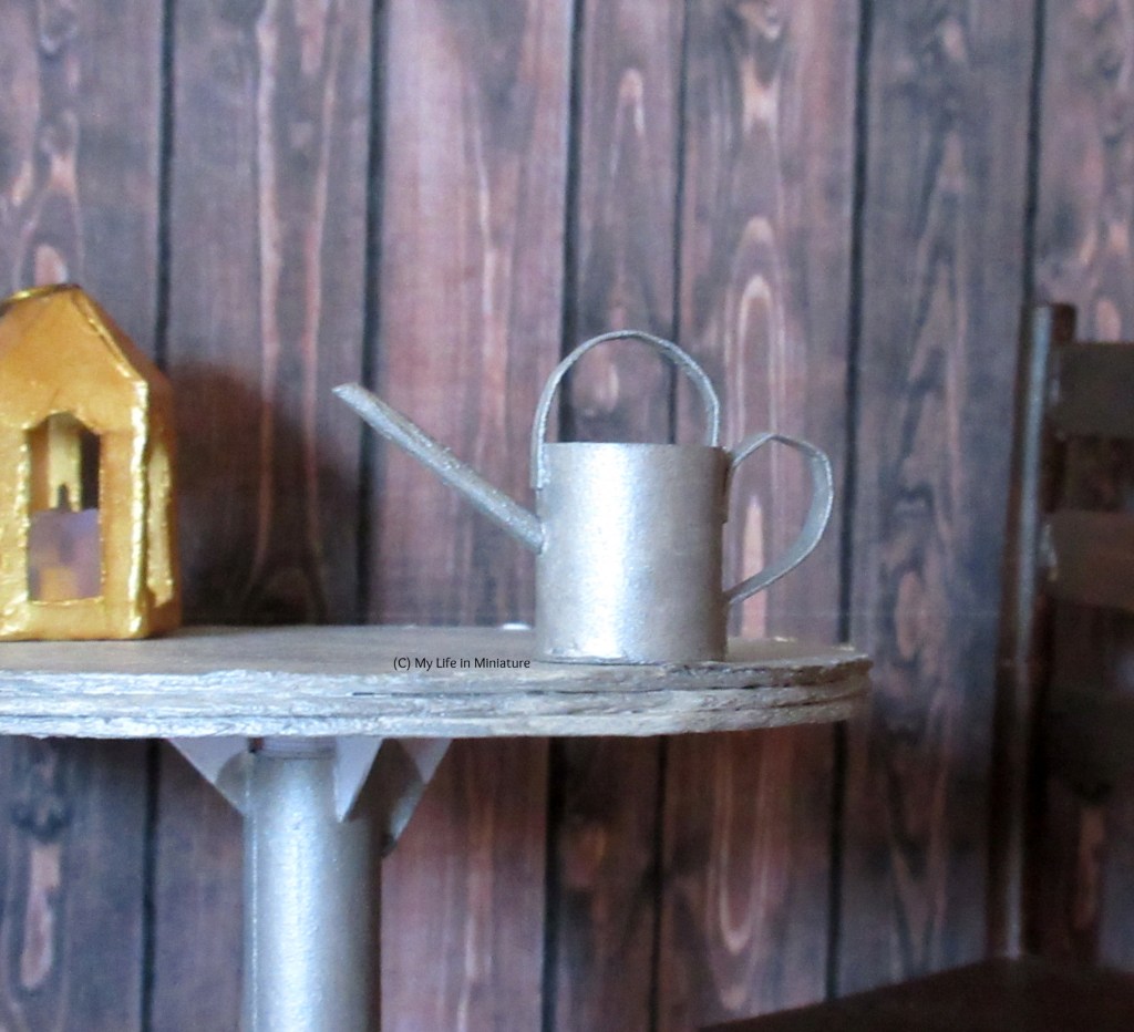 A silver watering can with a thin spout sits on the table in Tea at Yaz's courtyard. It has two handles: one on top and one on the side. 
