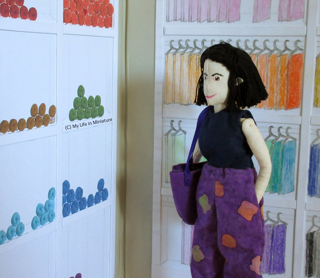 Tiffany stands in front of the two shelving units, looking at the yarn shelves. She has a hand in her pocket, a dark purple tote bag on her shoulder and a thoughtful expression.