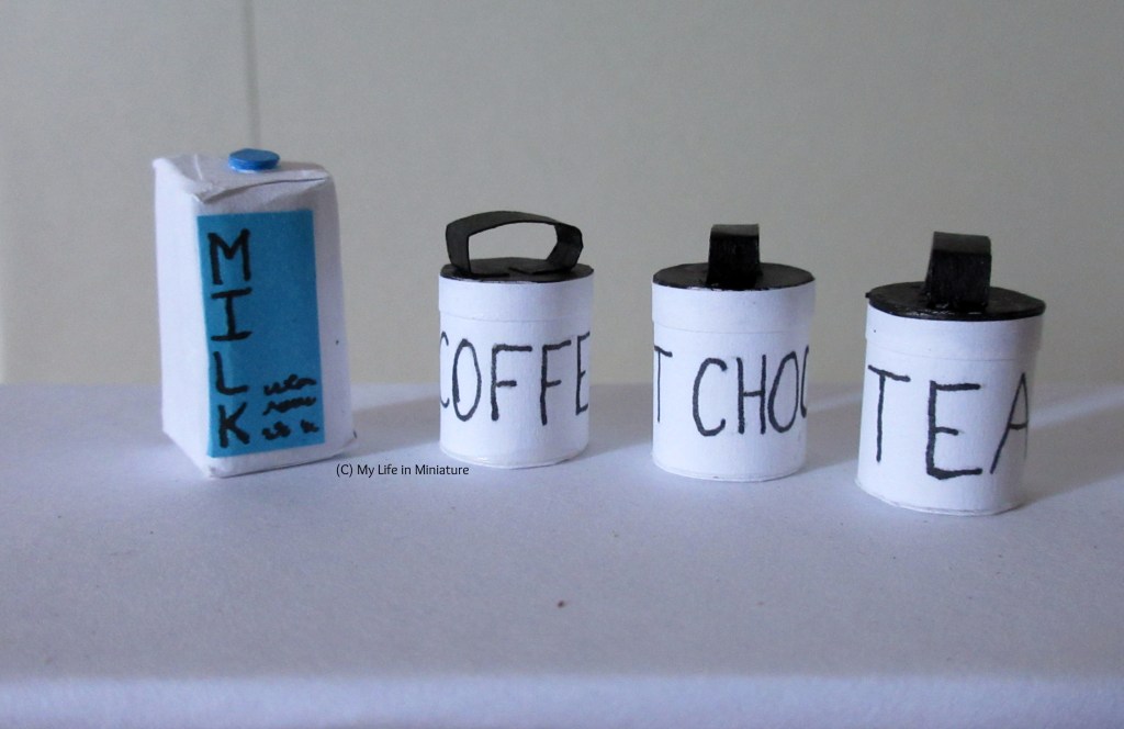The three canisters and the milks it on a white surface. The milk carton is white with a blue label saying 'MILK' and a blue screw-on lid on top. The three canisters are white with black lids and their contents written on them in black. 