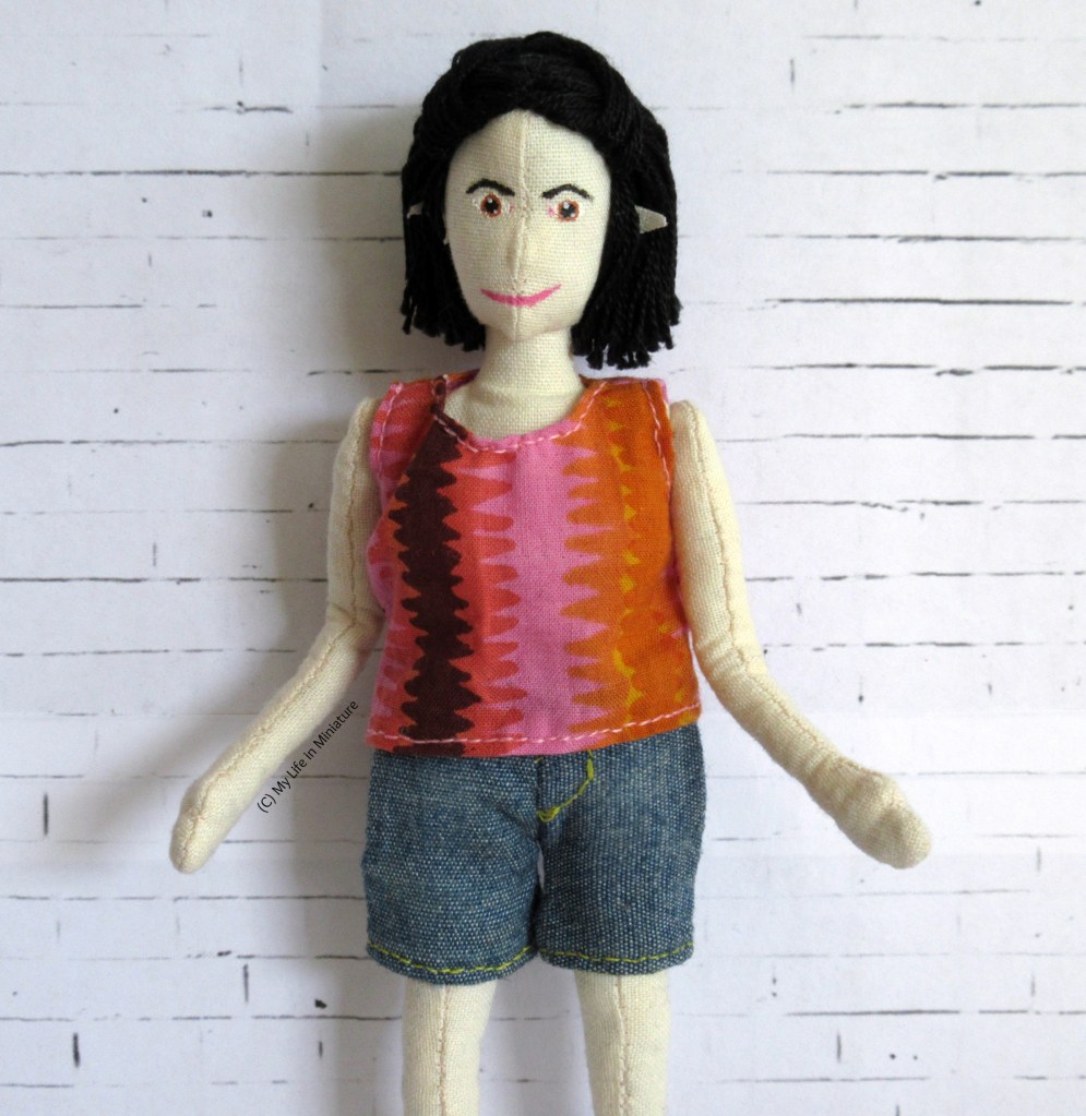 TIffany wears a pink, orange, red 
and dark brown shirt and denim shorts in front of a white brick background. Her hands are spread in a 'ta-daa!' gesture. 