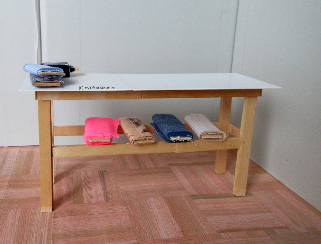 A table, with pale wood legs and a shiny white top, stands against a grey background and on wood parquet flooring. There are three small bolts of fabric on top, and four big bolts underneath. 
