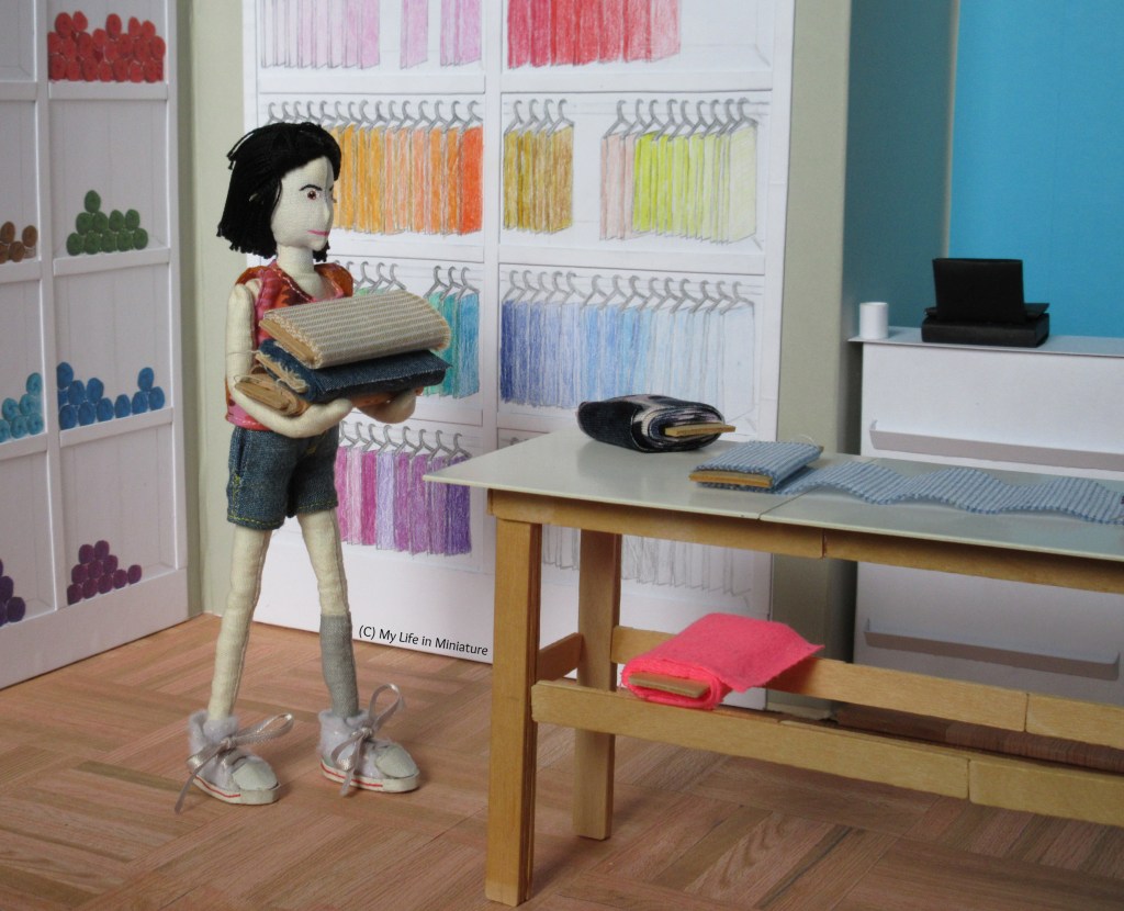 Tiffany carries three large bolts of fabric to the table, which sits in front of the counter and the shelving units inside Needle & Thread. A bolt of fabric is unwound on the table. 