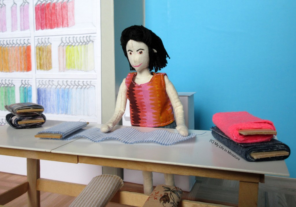 Tiffany stands at the table, bolts of fabric on wither side of her. Another bolt is unwound in front of her, and she is smoothing it out. 