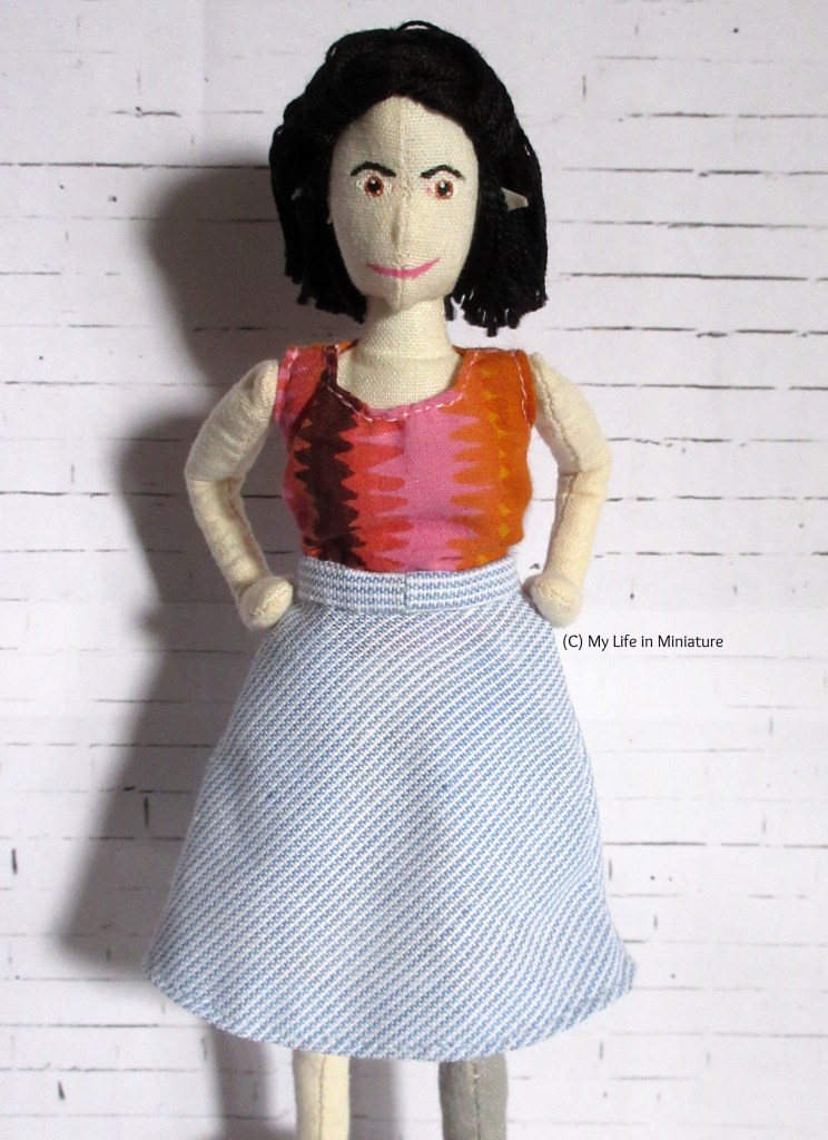 Tiffany stands facing the camera, wearing a white and blue pin-striped A-line skirt. Her hands are on her hips, and she is against a white brick background.