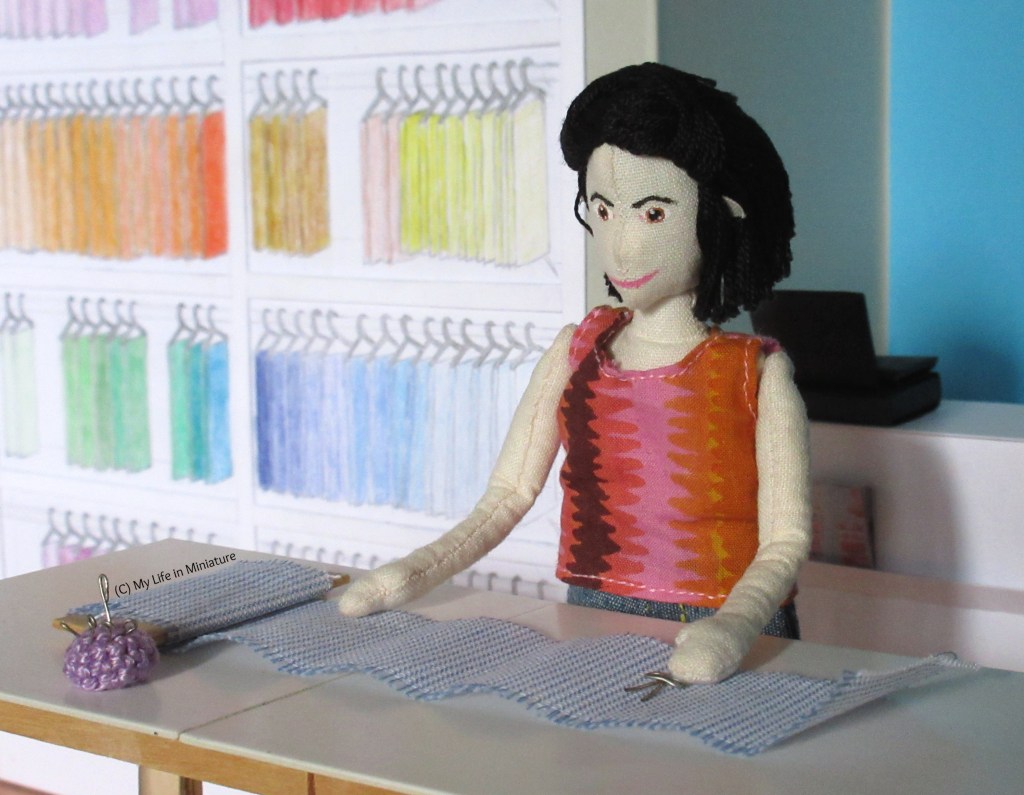 Tiffany stands at the fabric table in Needle & Thread, smoothing out a bolt of white and blue pinstriped fabric. One hand reaches for fabric scissors, ready to cut out pattern pieces. 