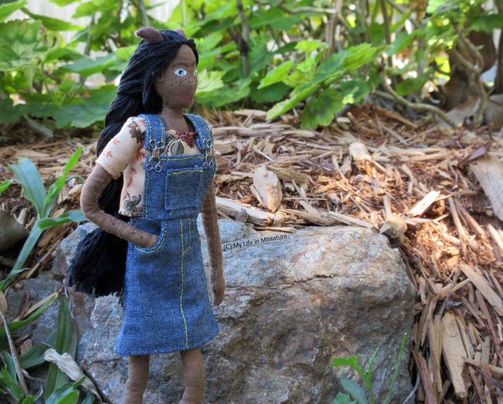 Petra stands beside a rock, a mulched garden bed behind her. She looks to the right and has a hand in the pocket of her skirt. 