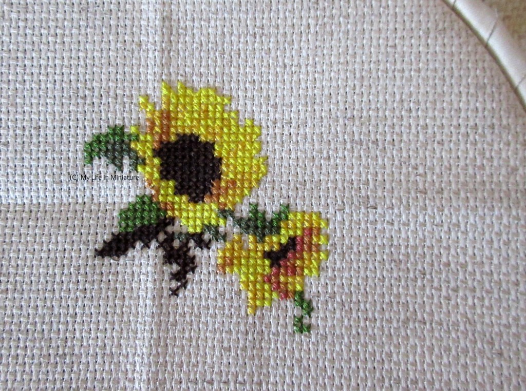Two sunflowers, with a bit more greenery around them, are cross-stitched onto cream fabric held in an embroidery hoop.
