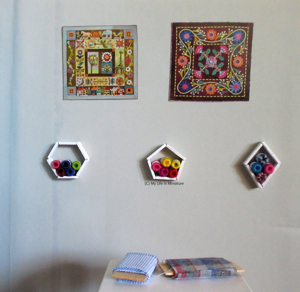 The grey wall beside the fabric table is facing the camera, with the fabric table visible in the foreground. On the wall, there are two images of quilts, and below that is three white shelves stacked with yarn balls.