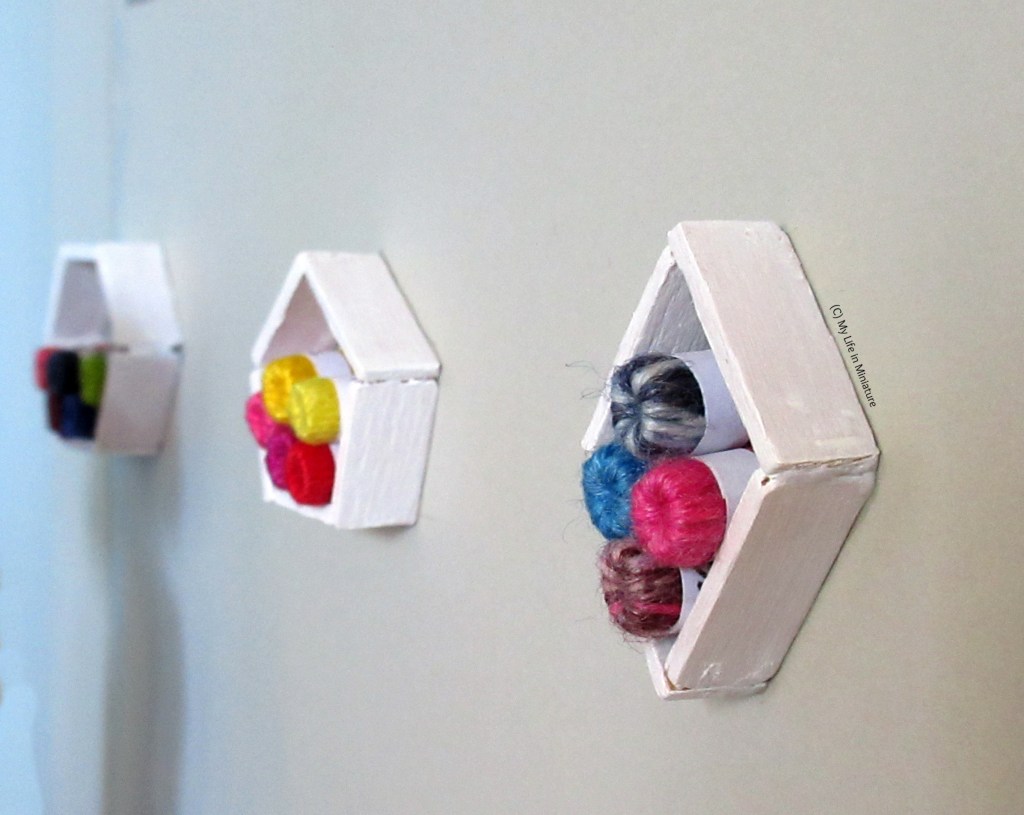 Close-up of one of the shelves, with the other two visible but blurry in the background. Closest shelf is diamond-shaped, next closest is a pentagon, farthest is a hexagon. All are painted white and stacked with balls of yarn.