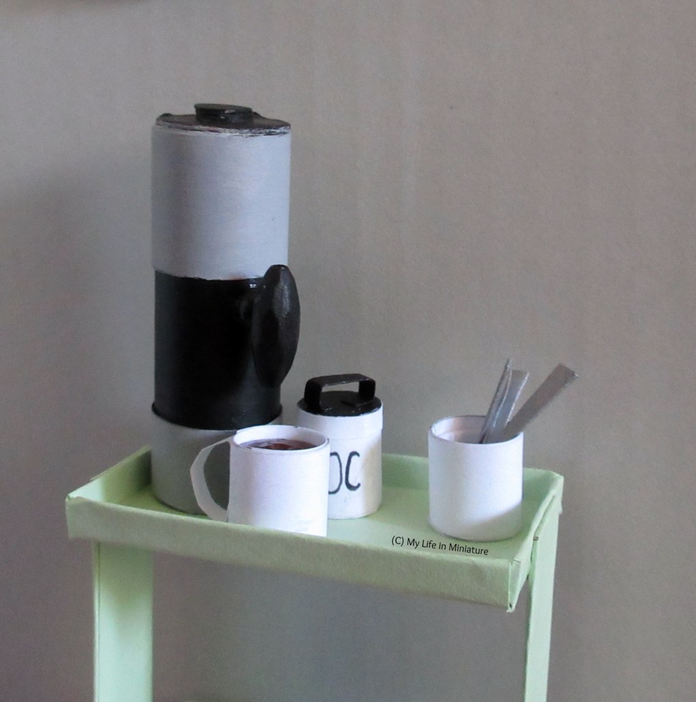 The top tray of the drinks trolley is against a grey background. A white food canister, like a porcelain cookie jar without a lid, sits on top holding silver teaspoons. Also on top is the hot water urn, a mug of hot chocolate, and the white hot chocolate jar.