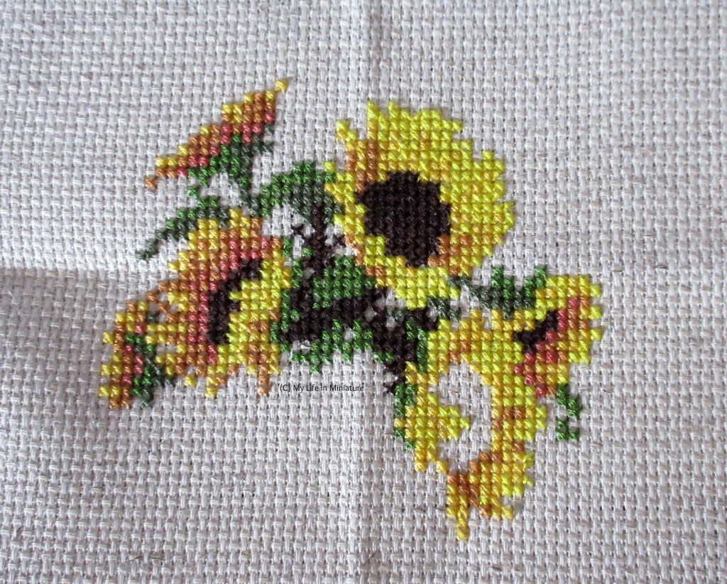 Five-and-a-half sunflowers, and some greenery and stems, are cross-stitched onto cream fabric. 