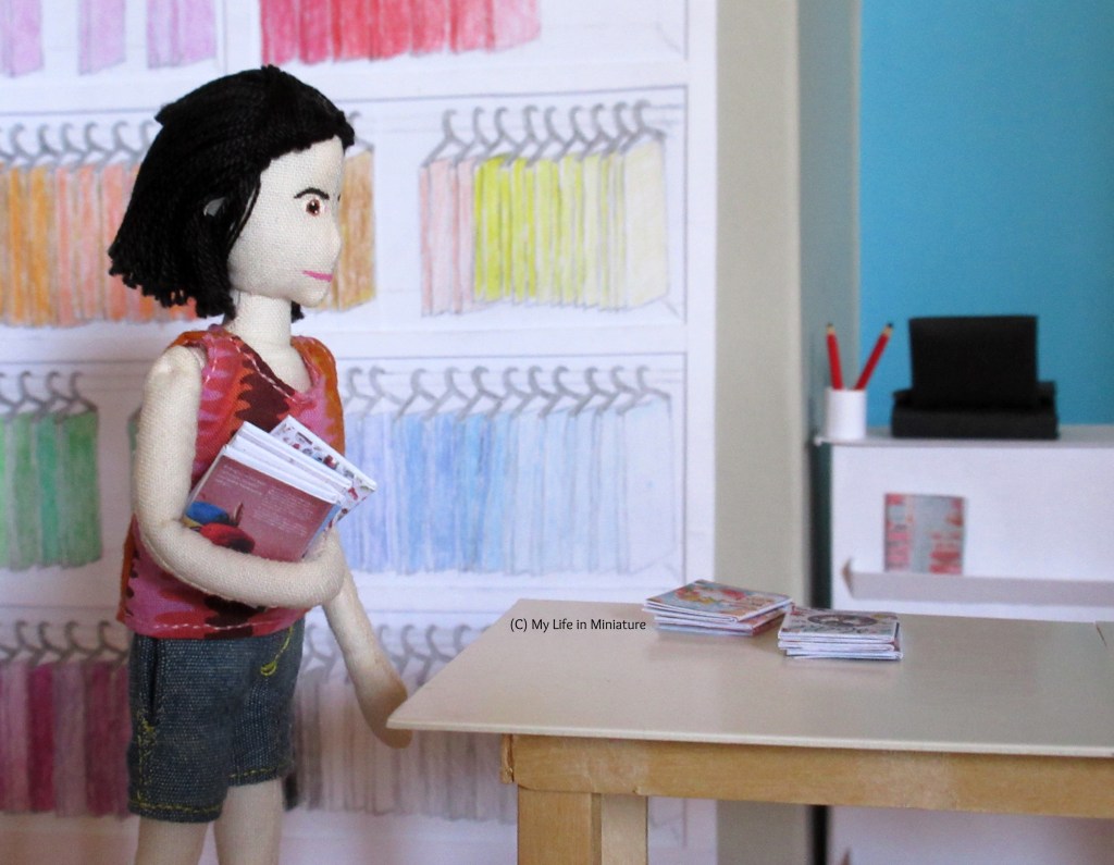 Tiffany carries an armful of books to the fabric table in Needle & Thread. Two piles of magazines sit on the fabric table, having already been unpacked.